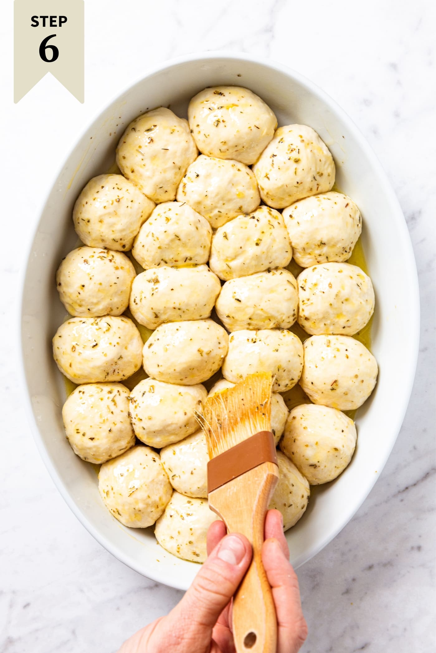 Hand using a food brush to brush melted butter and Italian seasoning mixture over the top of unbaked pizza bites in white oval baking dish sitting on marble surface.