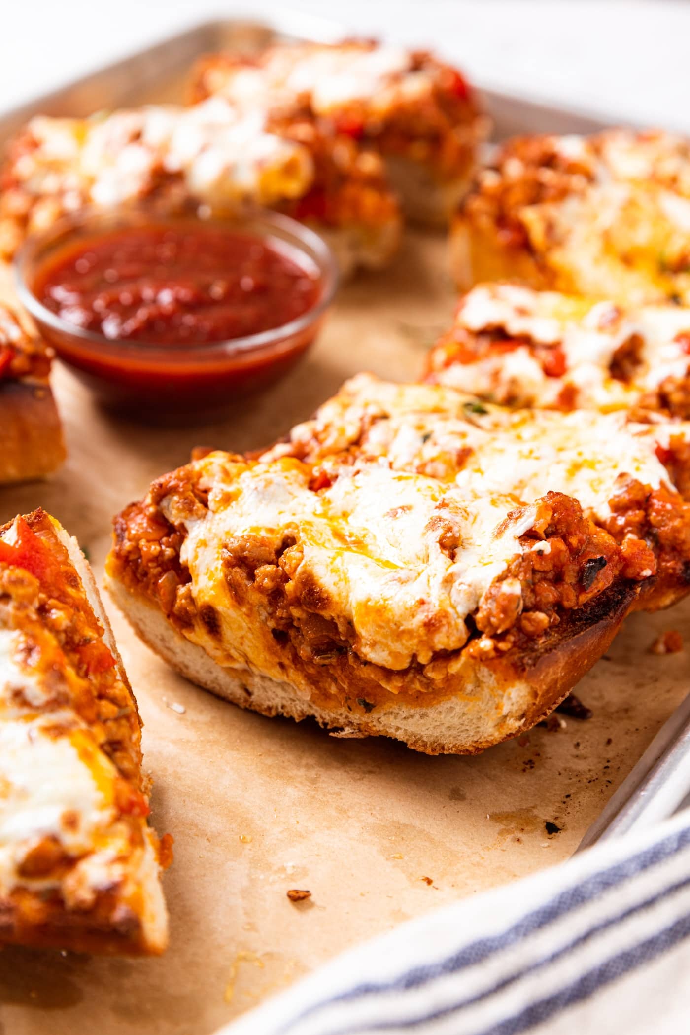 Slice of French bread pizza topped with tomato and sausage sauce with melted cheese sitting on parchment-lined baking sheet with small glass bowl filled with dipping sauce.