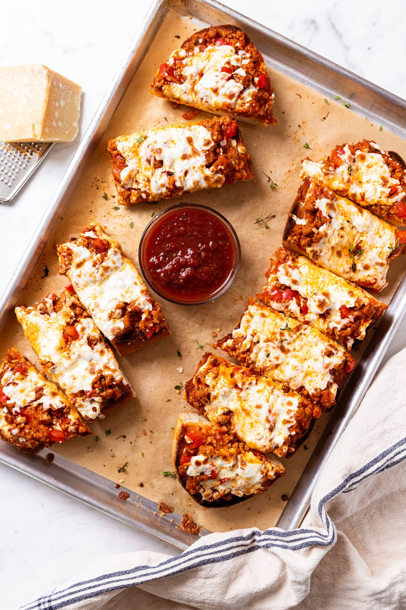 Baking sheet with piece of brown-colored parchment paper sitting on white surface filled with slices of sausage French bread pizza with small glass bowl of dipping sauce.