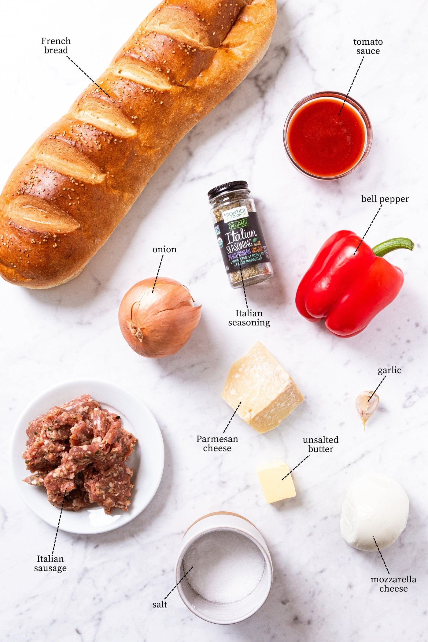 White marble surface filled with ingredients needed to make French bread pizza including a loaf of French bread, bell pepper, onion, sausage, salt, cheese, and more.
