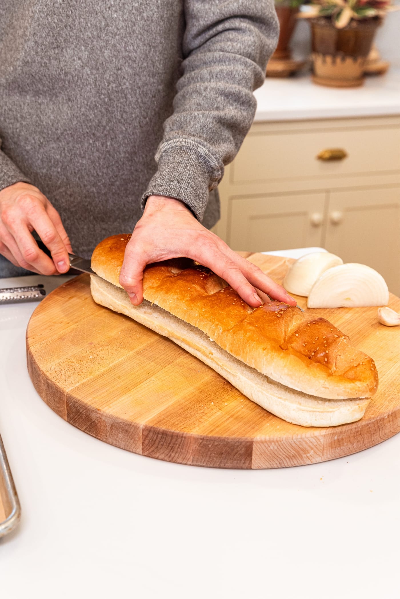 Hand holding onto the top of a loaf of French bread slicing it in half on a wood cutting board with halved onion in background.
