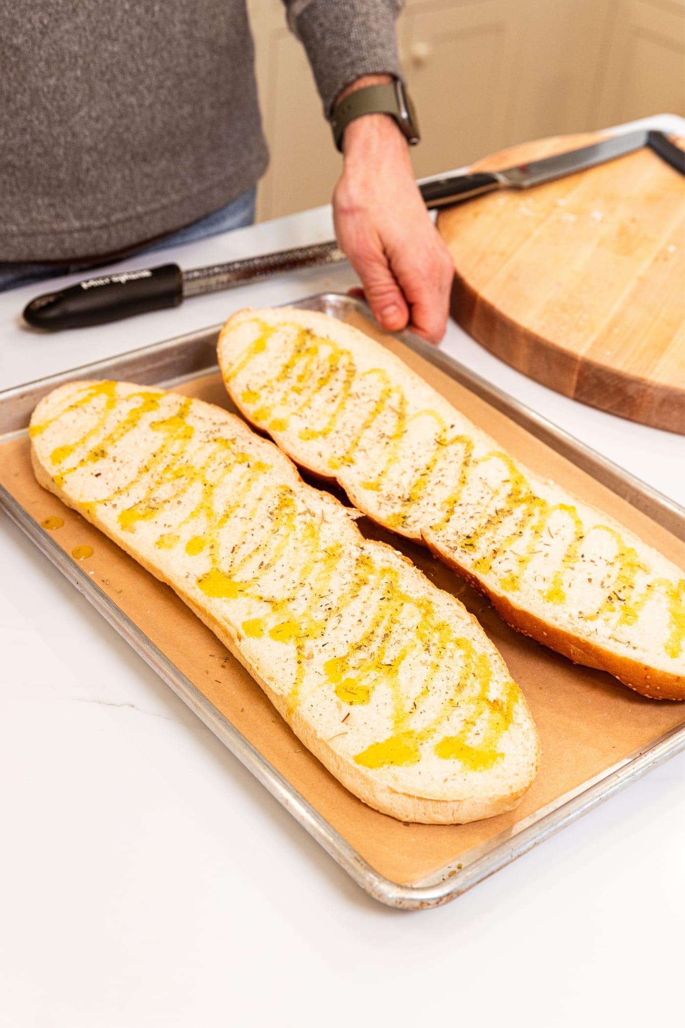 Two halves of French bread with inside showing drizzled with olive oil, black pepper, and salt sitting on a parchment-lined baking sheet on white surface.