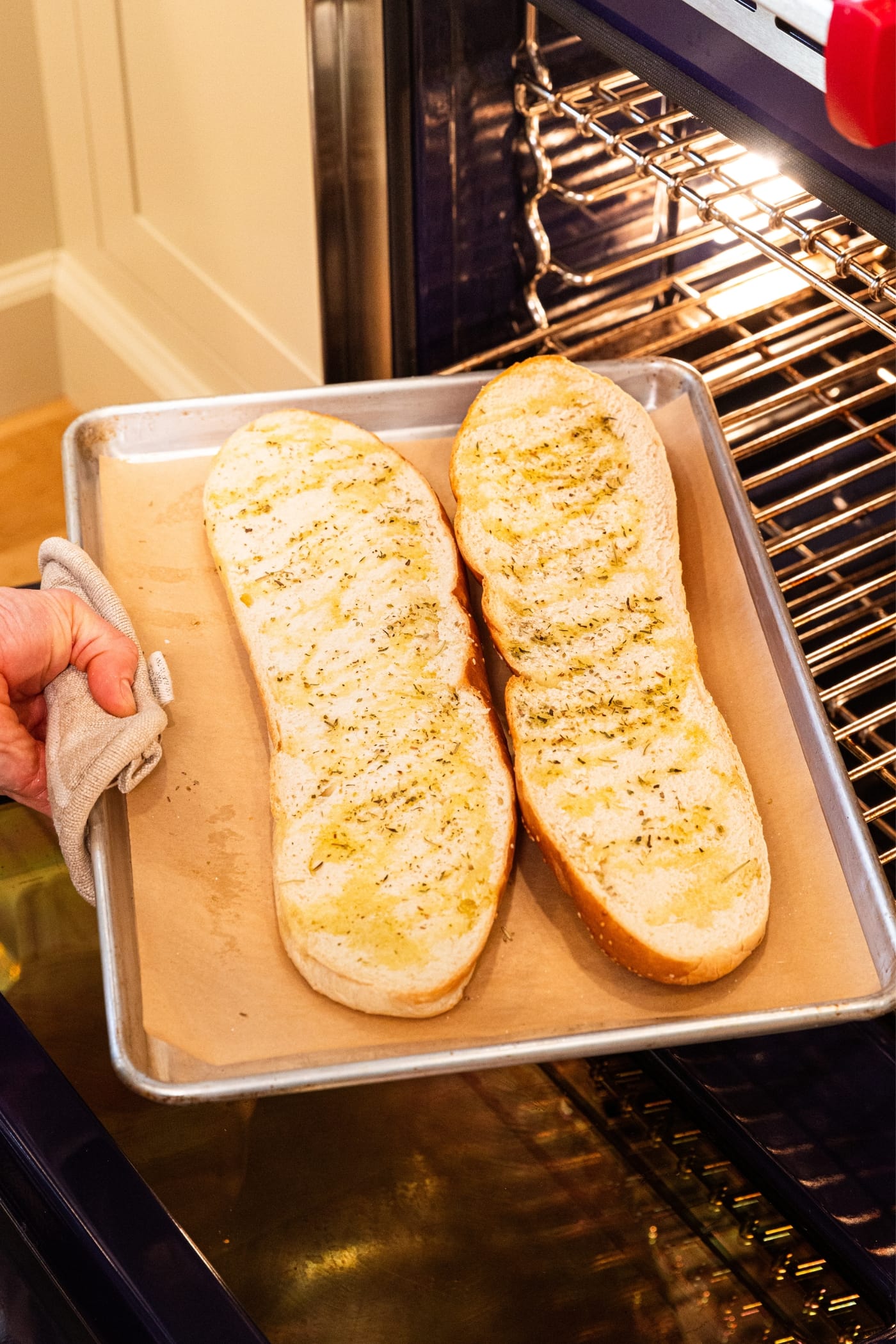 Hand holding onto parchment-lined baking sheet pulling crisped halves of French bread out of a hot oven.