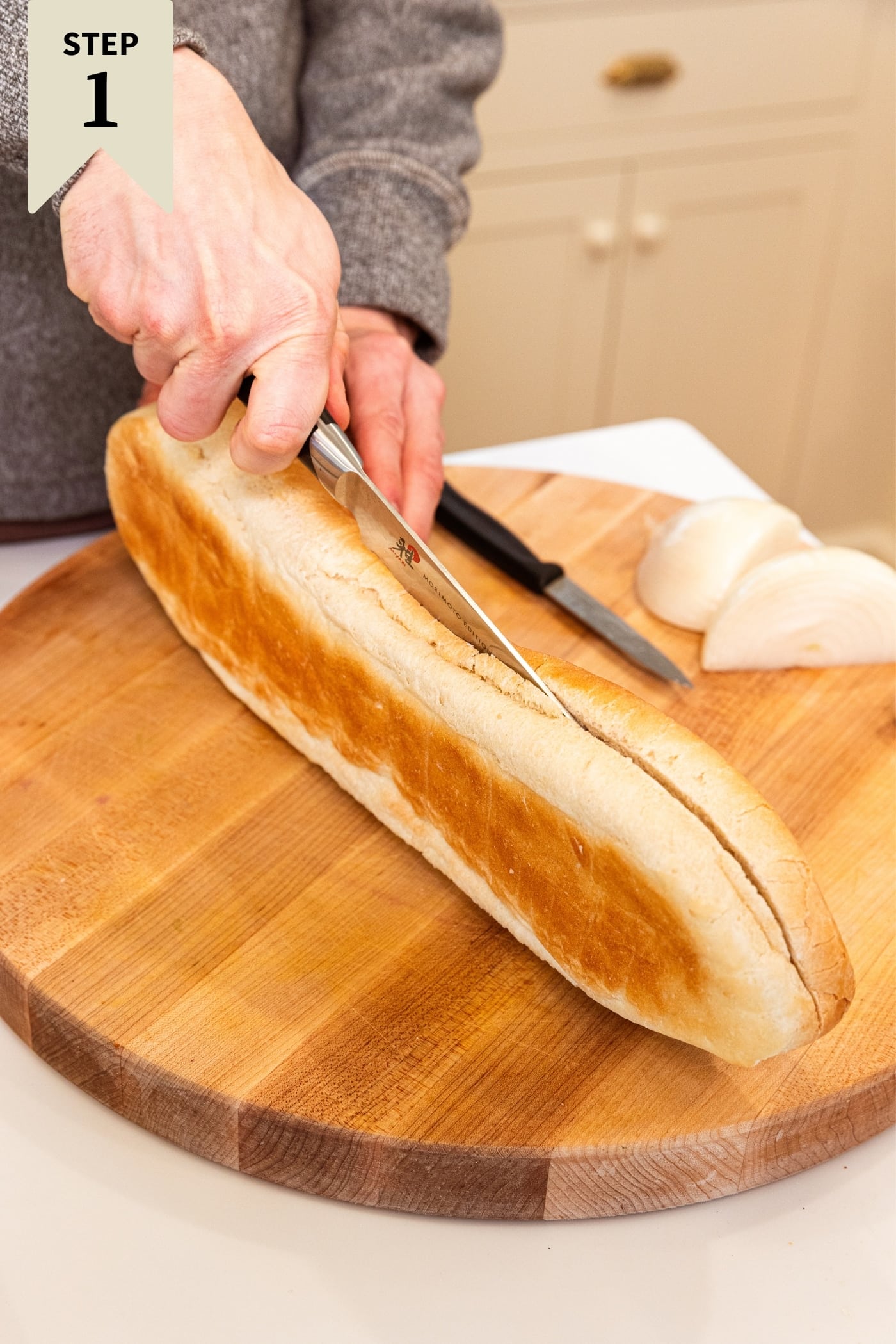 Hand using knife to slice open a browned loaf of French bread on a wood cutting board with chopped onion to the side.