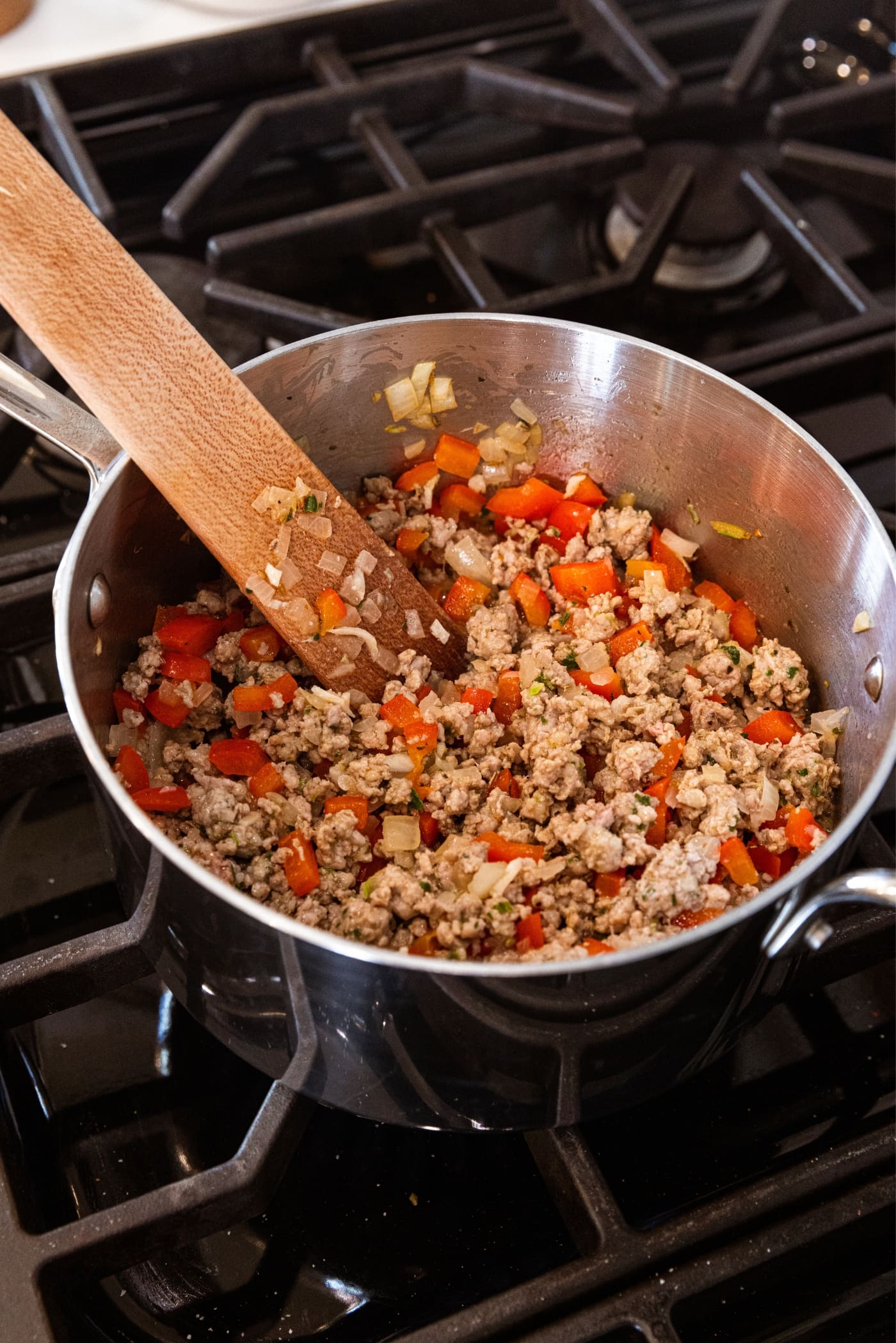 Wooden spoon resting against the side of metal saucepan sitting on stovetop filled with browned sausage and pieces of diced red bell pepper and onion.