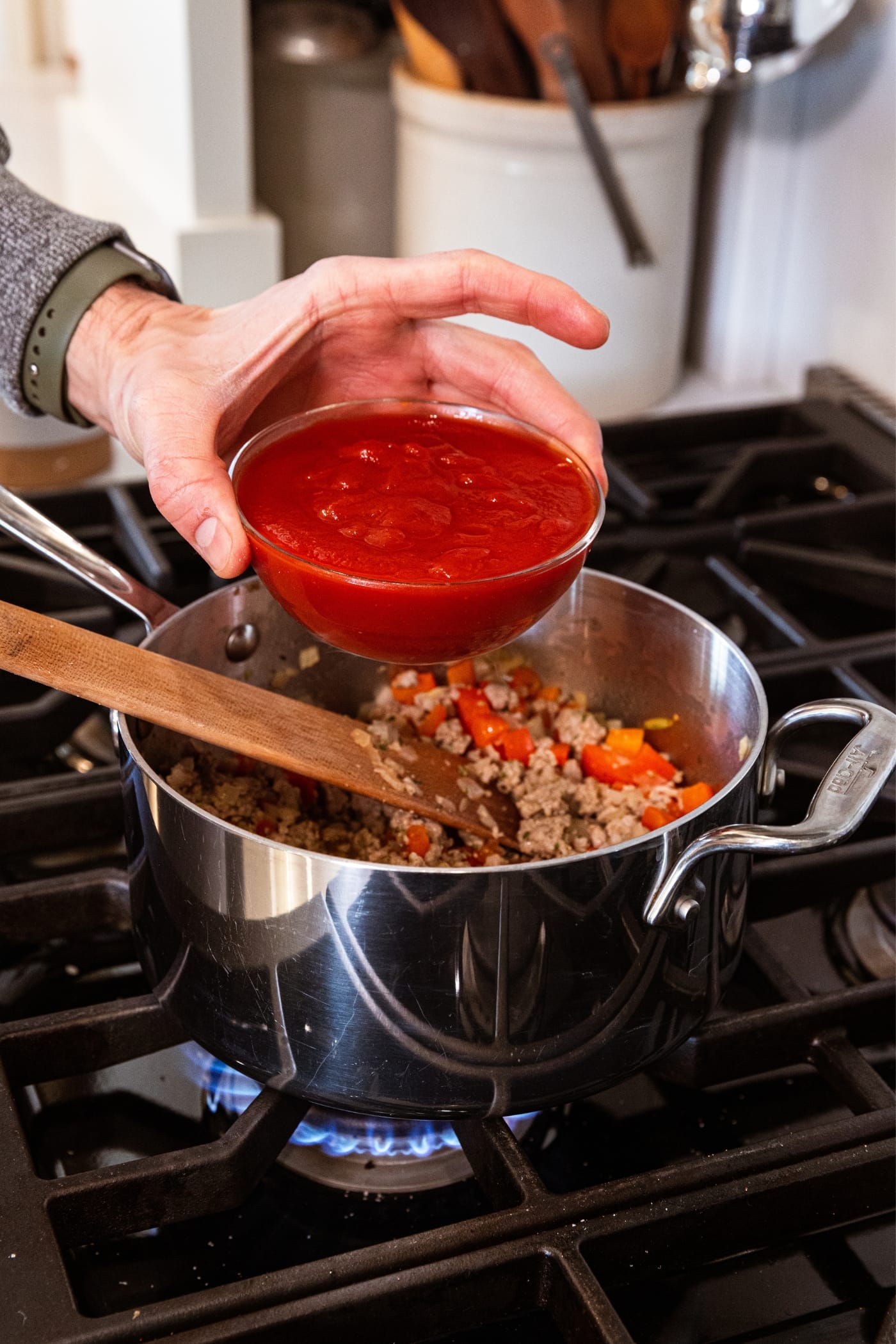 Hand holding tomato sauce in small glass bowl about to pour it into a metal saucepan filled with browned sausage and red bell pepper.