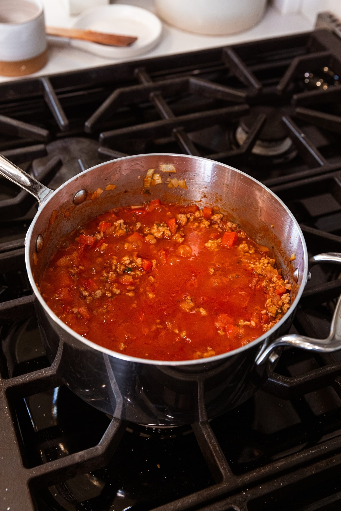 Metal saucepan on stovetop filled with homemade sausage tomato sauce with bits of sausage and tomato liquid showing along with small pieces of red bell pepper.
