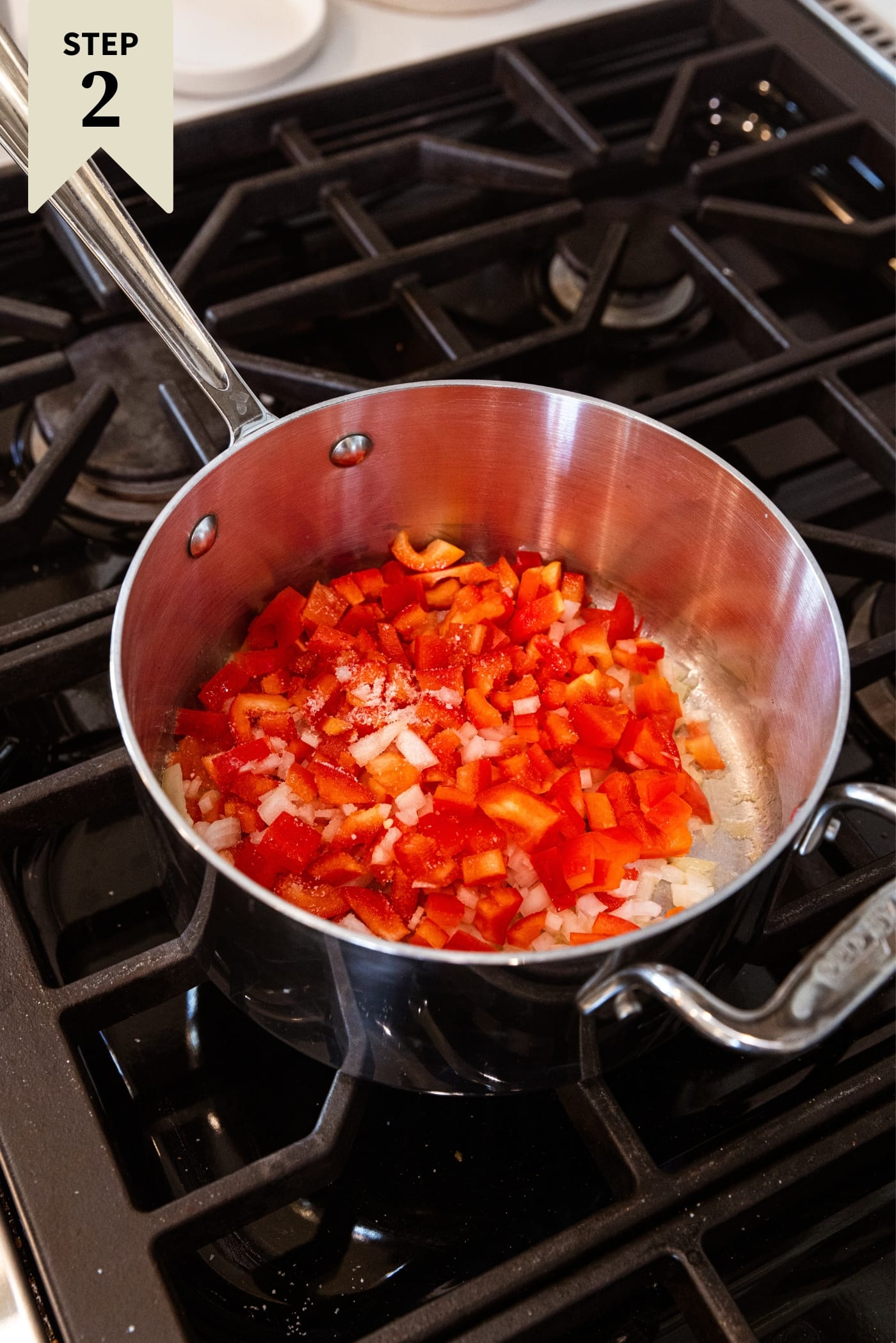 Metal saucepan sitting on stovetop with raw red bell pepper and chopped onion before being browned.