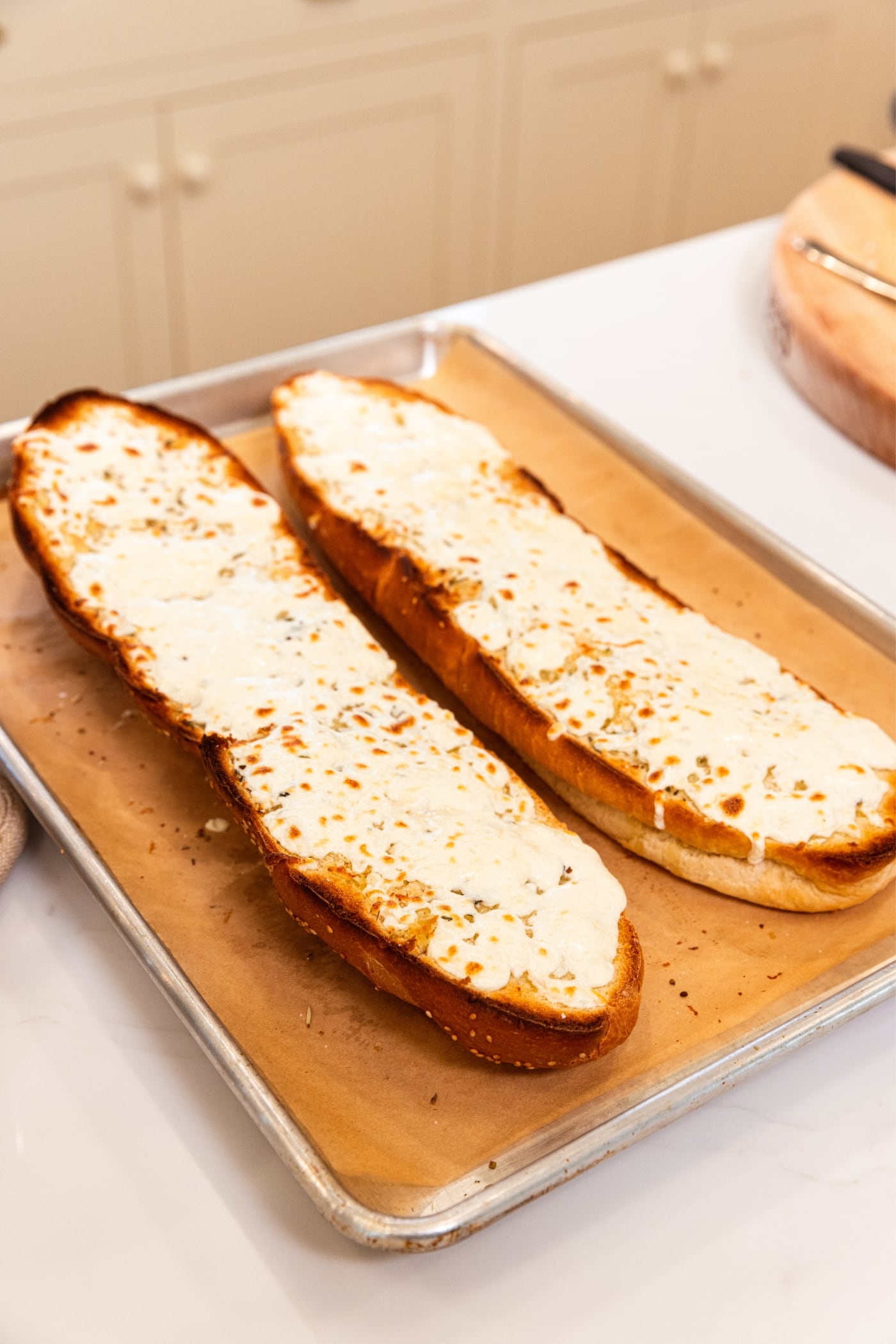 Two halves of French bread sitting on a parchment-lined baking sheet with melted cheese all over the top right after coming out of the oven.