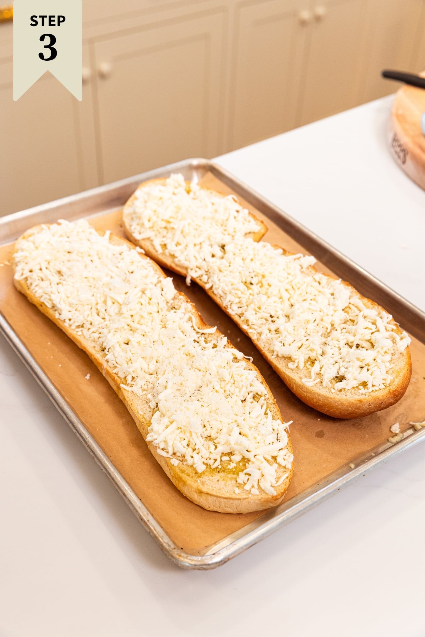 White surface with parchment-lined baking sheet filled with untoasted loaves of French bread with shredded unmelted cheese on top right before going in the oven.