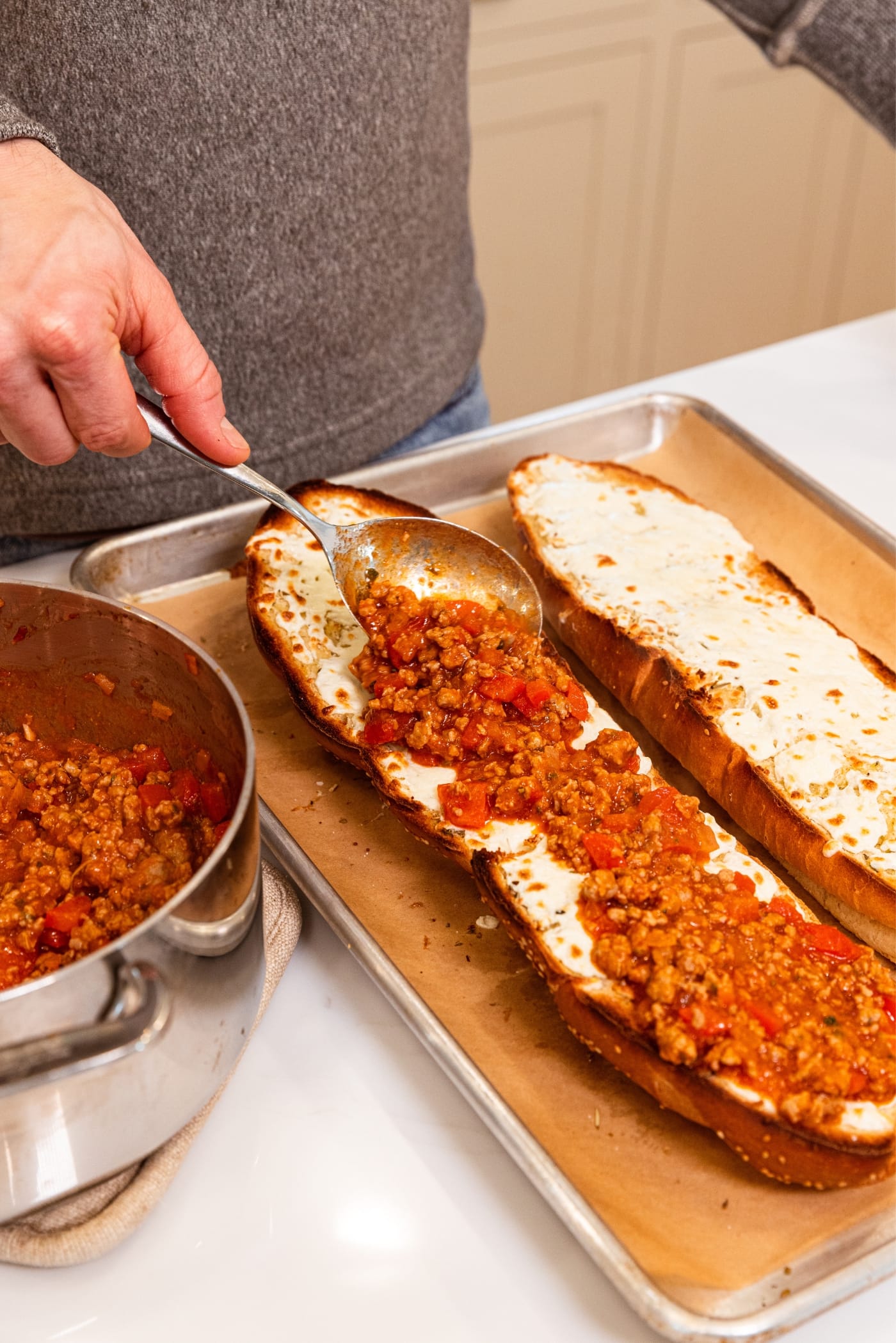 Hand holding spoon placing a spoonful of sausage meat mixture with tomato sauce on top of half of French bread topped with shredded and melted cheese.