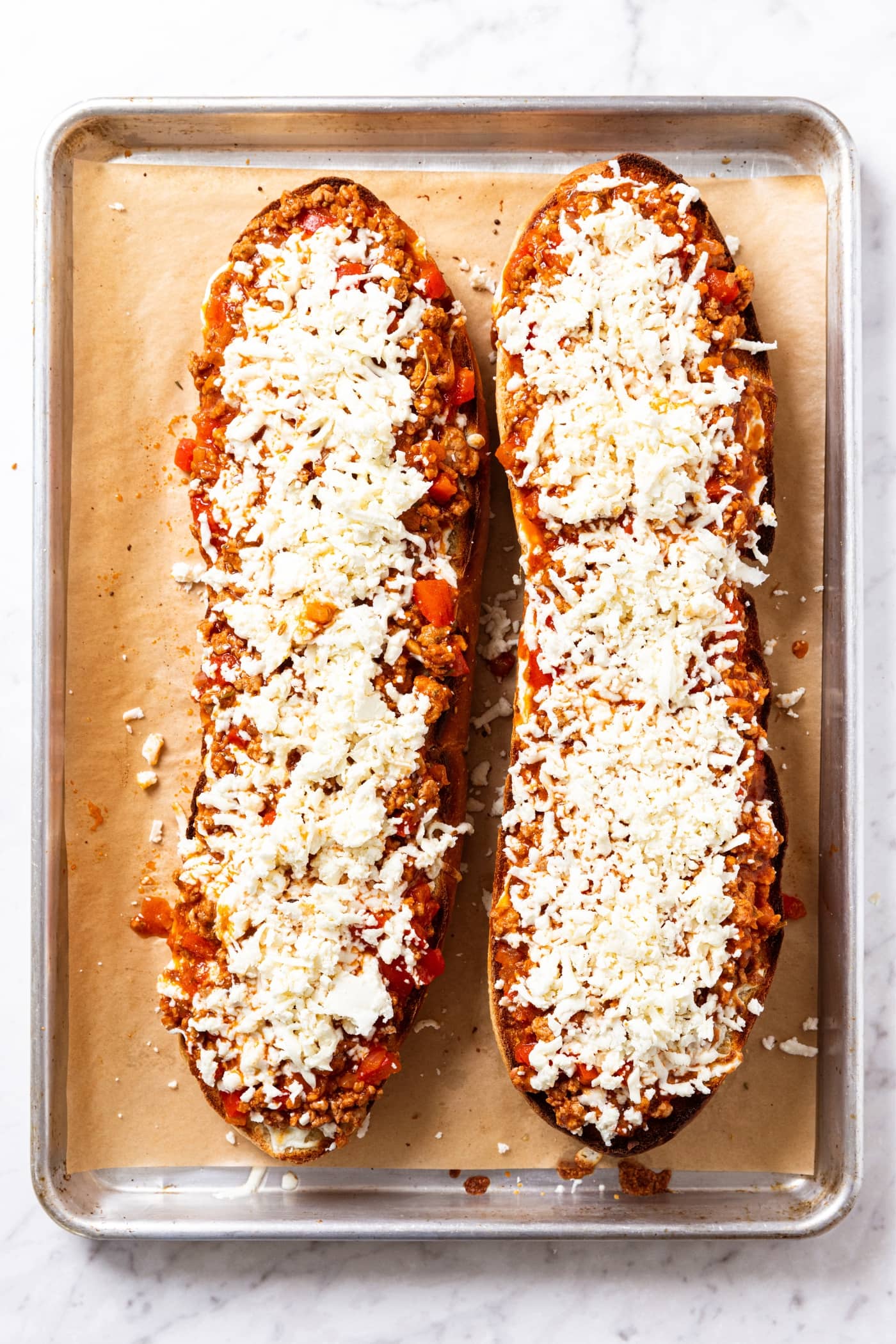 Two halves of French bread sitting on parchment-lined baking sheet sprinkled with shredded cheese right before going into the oven to bake.
