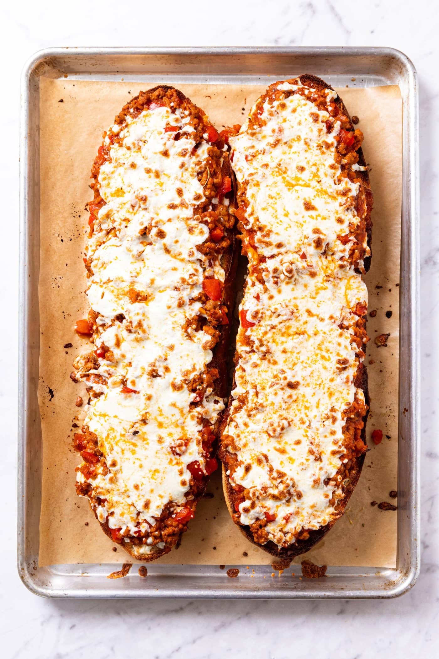 Baked halves of French bread pizza sitting on parchment-lined baking sheet with melted cheese all over the top right after coming out of the oven.