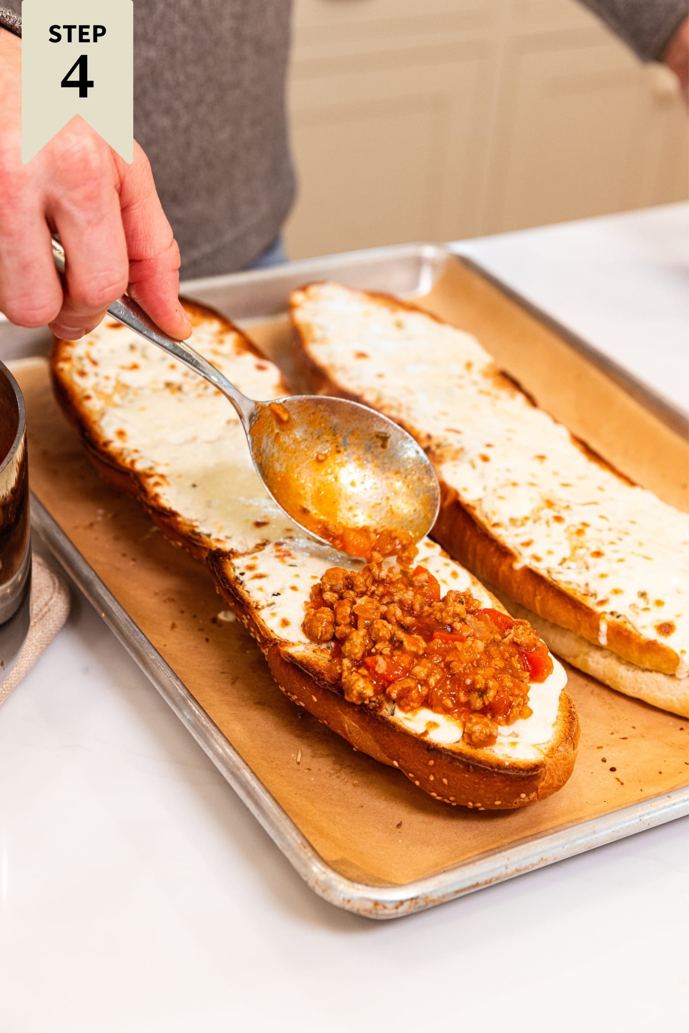 Hand holding spoon placing sausage tomato sauce mixture on top of cheese melted on French bread sitting on baking sheet on white surface.