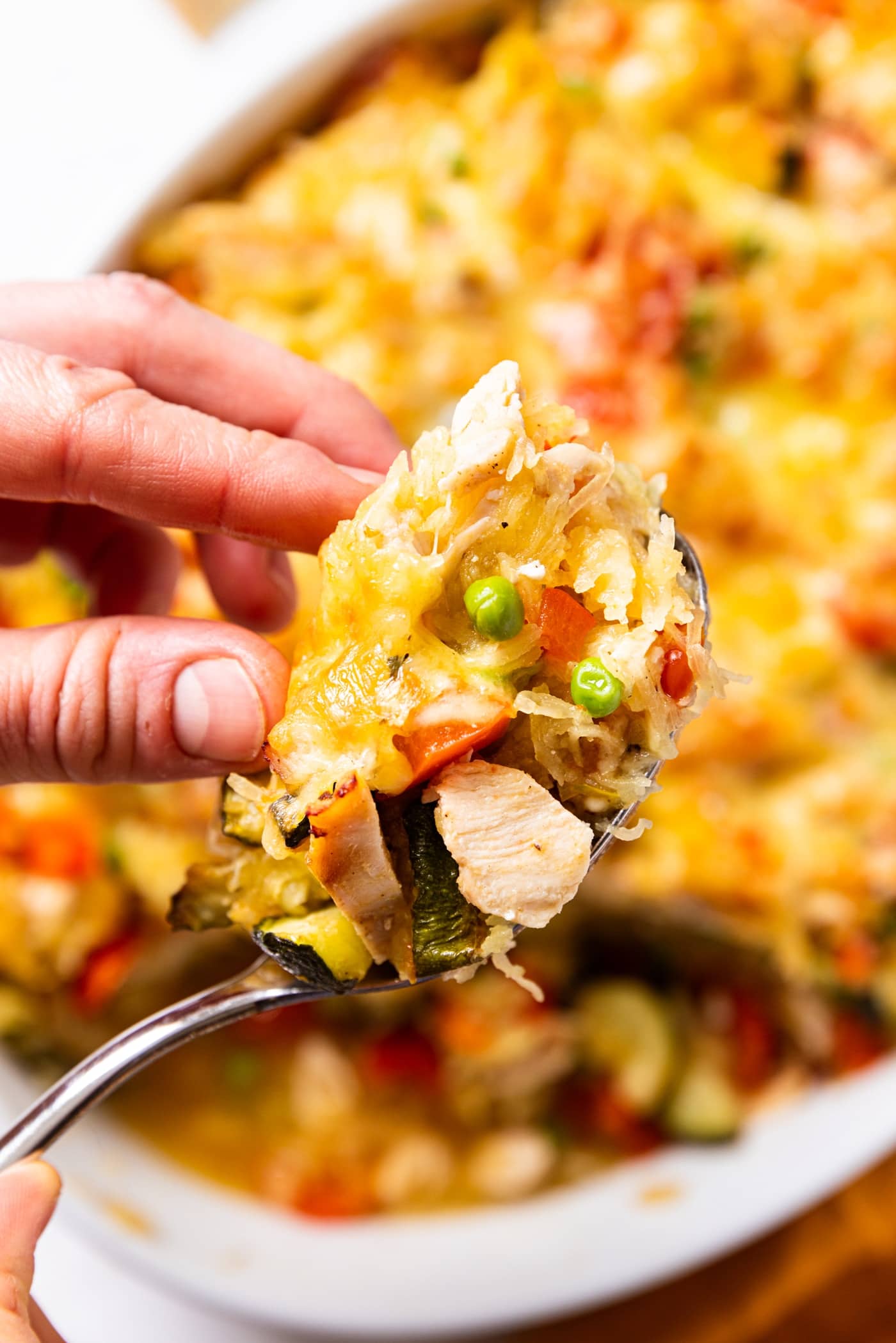 Hand holding spoonful of spaghetti squash chicken casserole with all ingredients showing including zucchini, peas, squash, and chicken.