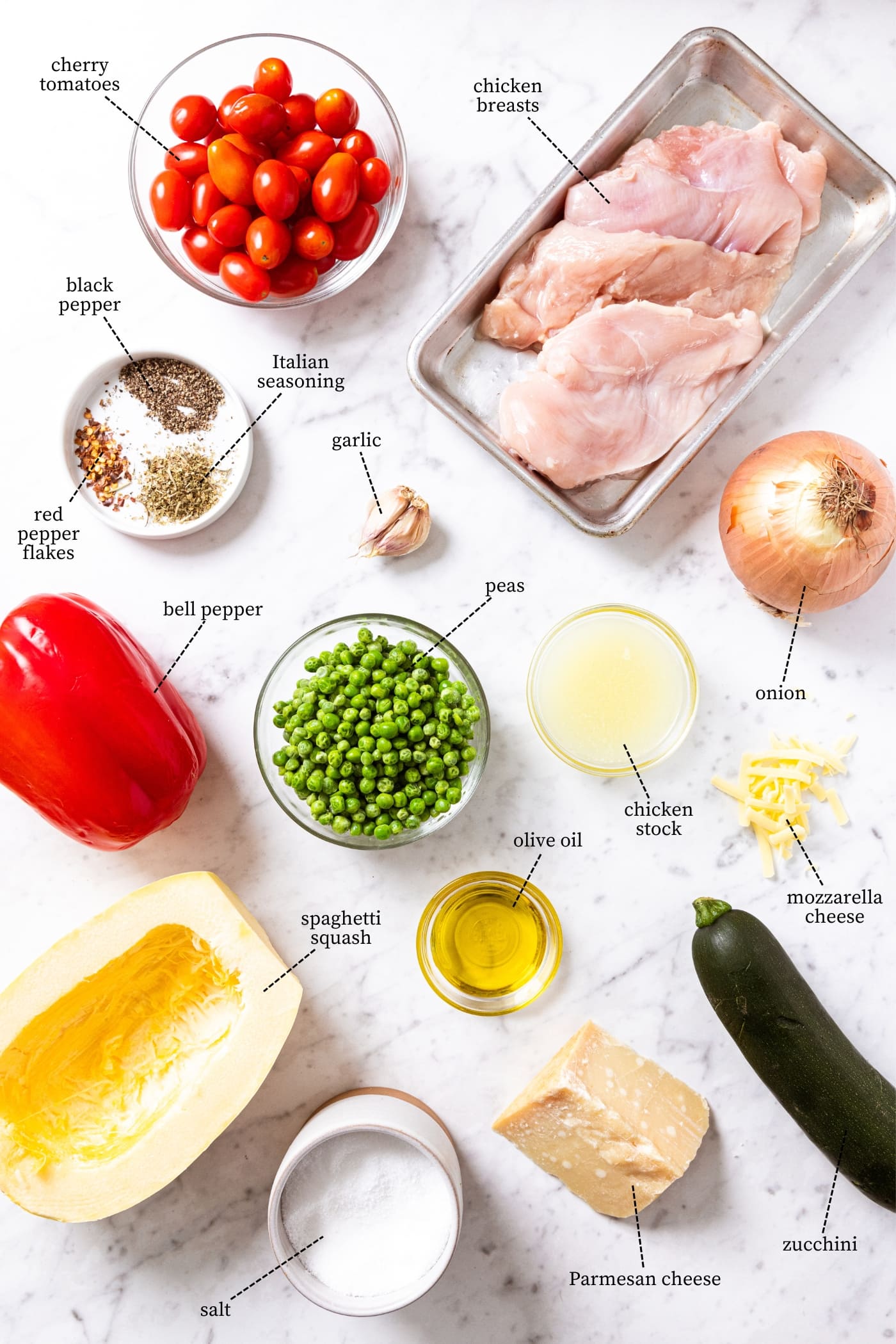 White marble surface filled with ingredients needed to make a casserole including spaghetti squash, bell pepper, peas, zucchini, onion, chicken, spices, and more.