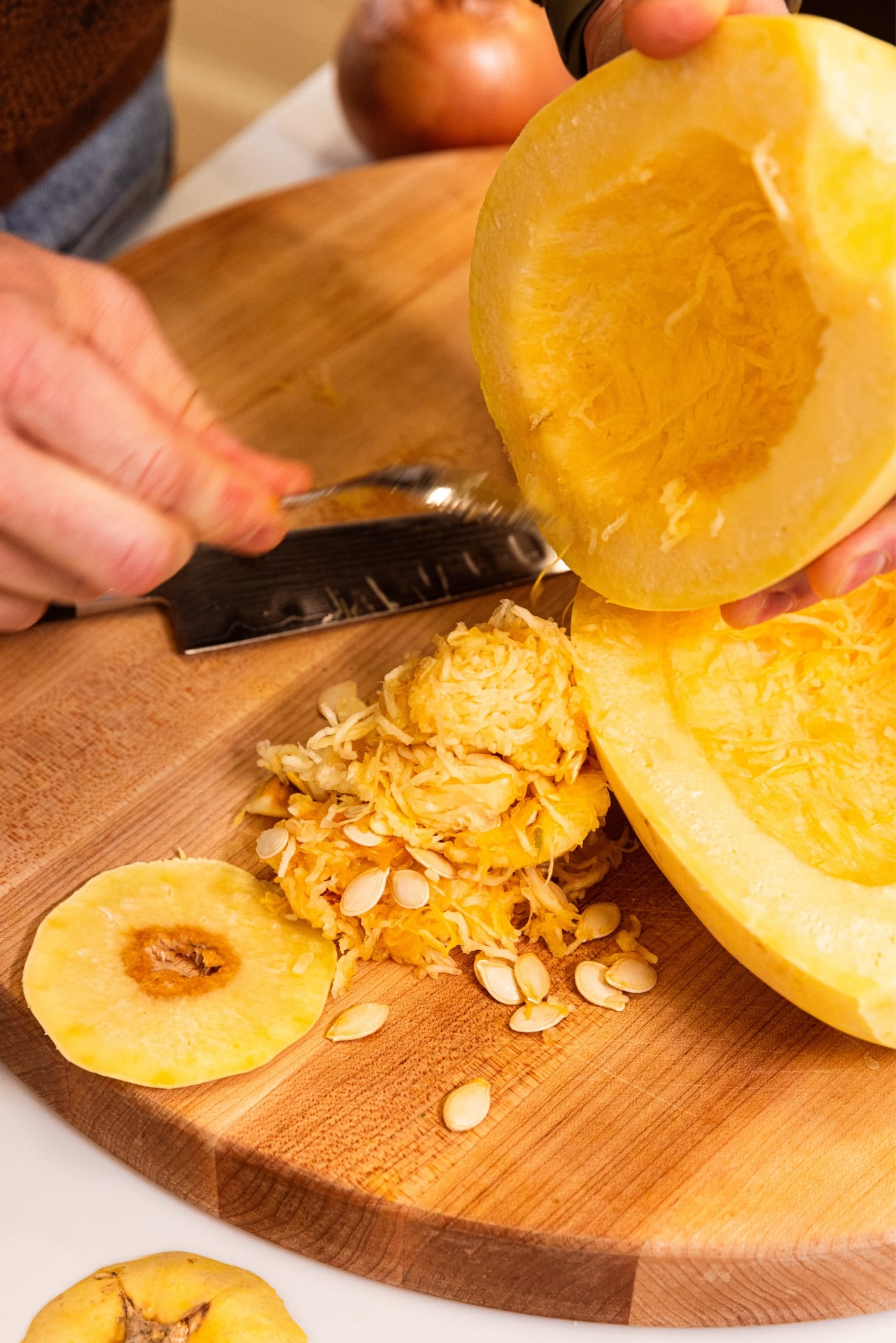 Hand using a spoon to scoop out the interior of a halved spaghetti squash onto a wooden cutting board.