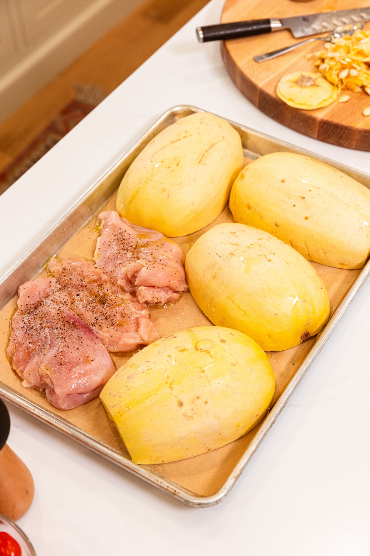 Large metal baking sheet with piece of parchment on bottom sitting on white surface with unbaked halves of spaghetti squash and raw chicken breasts with olive oil, salt, and pepper all over.
