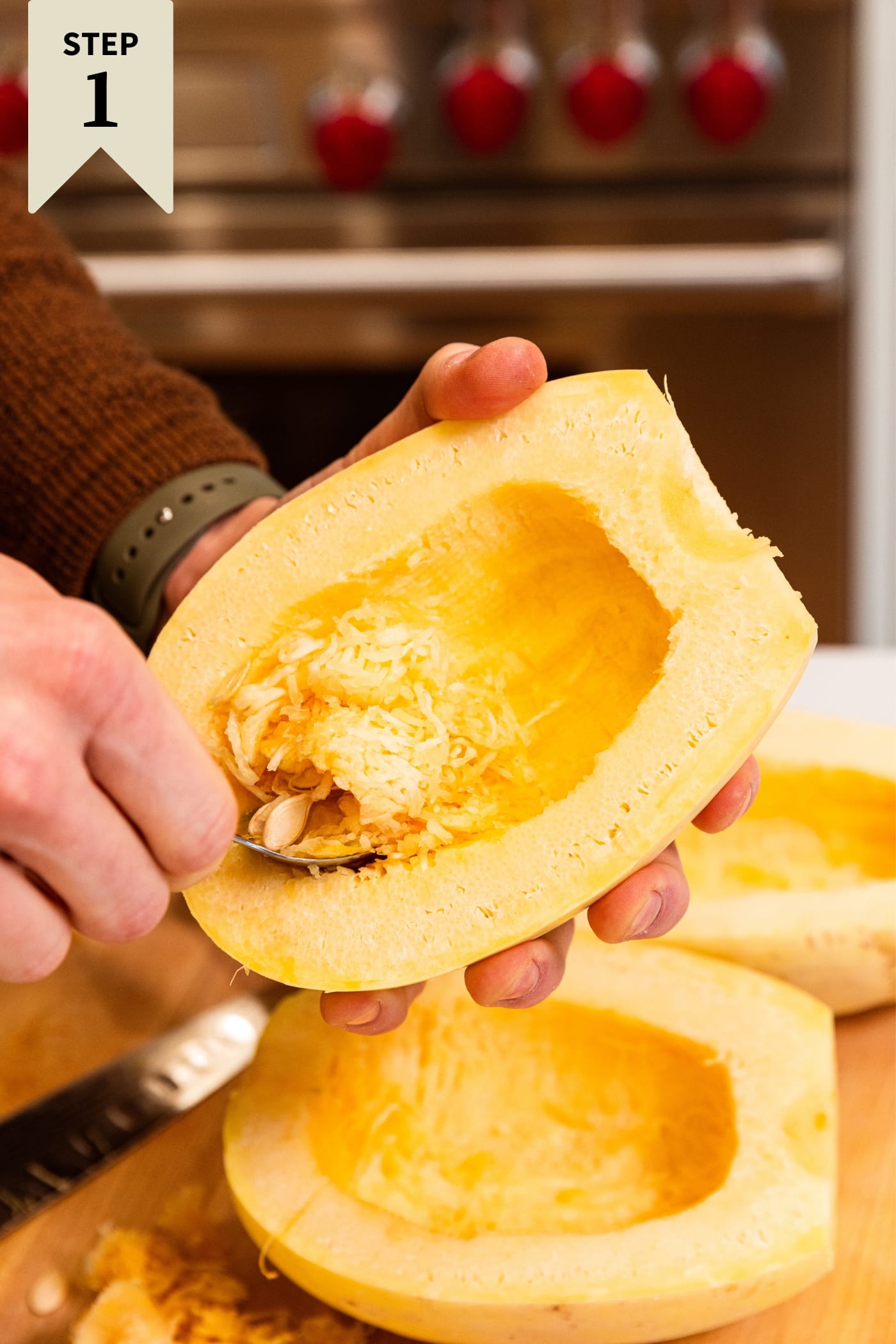 Hand holding spaghetti squash cut in half using a spoon to scoop out the interior flesh and seeds with more squash in background.