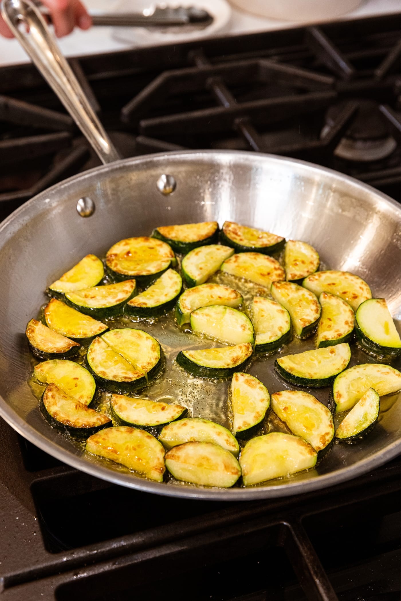 Browned half moons of zucchini sitting in metal skillet filled with oil over hot heat on stovetop.