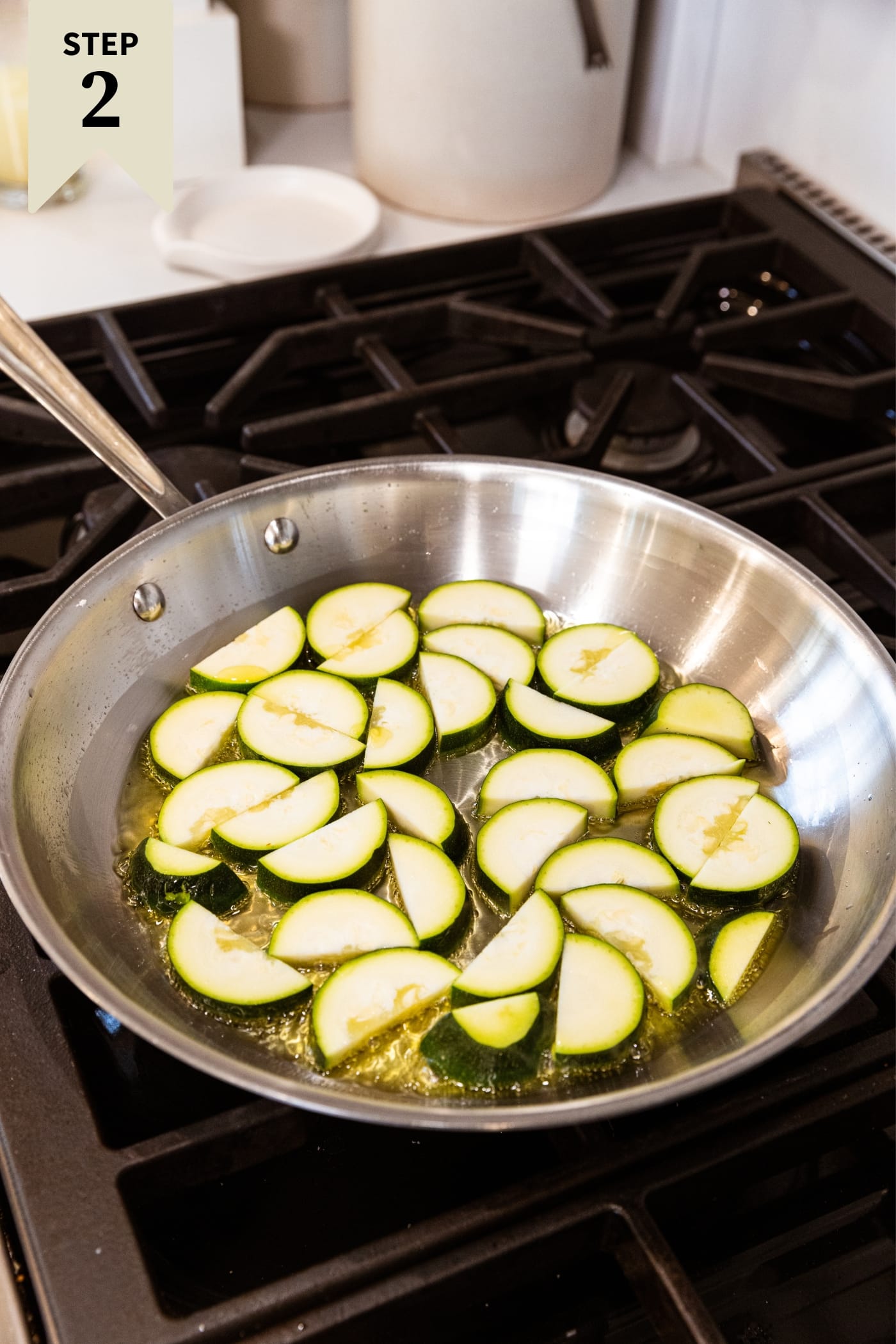 Metal skillet sitting on stovetop with uncooked half moons of zucchini sitting in hot oil on the heat.