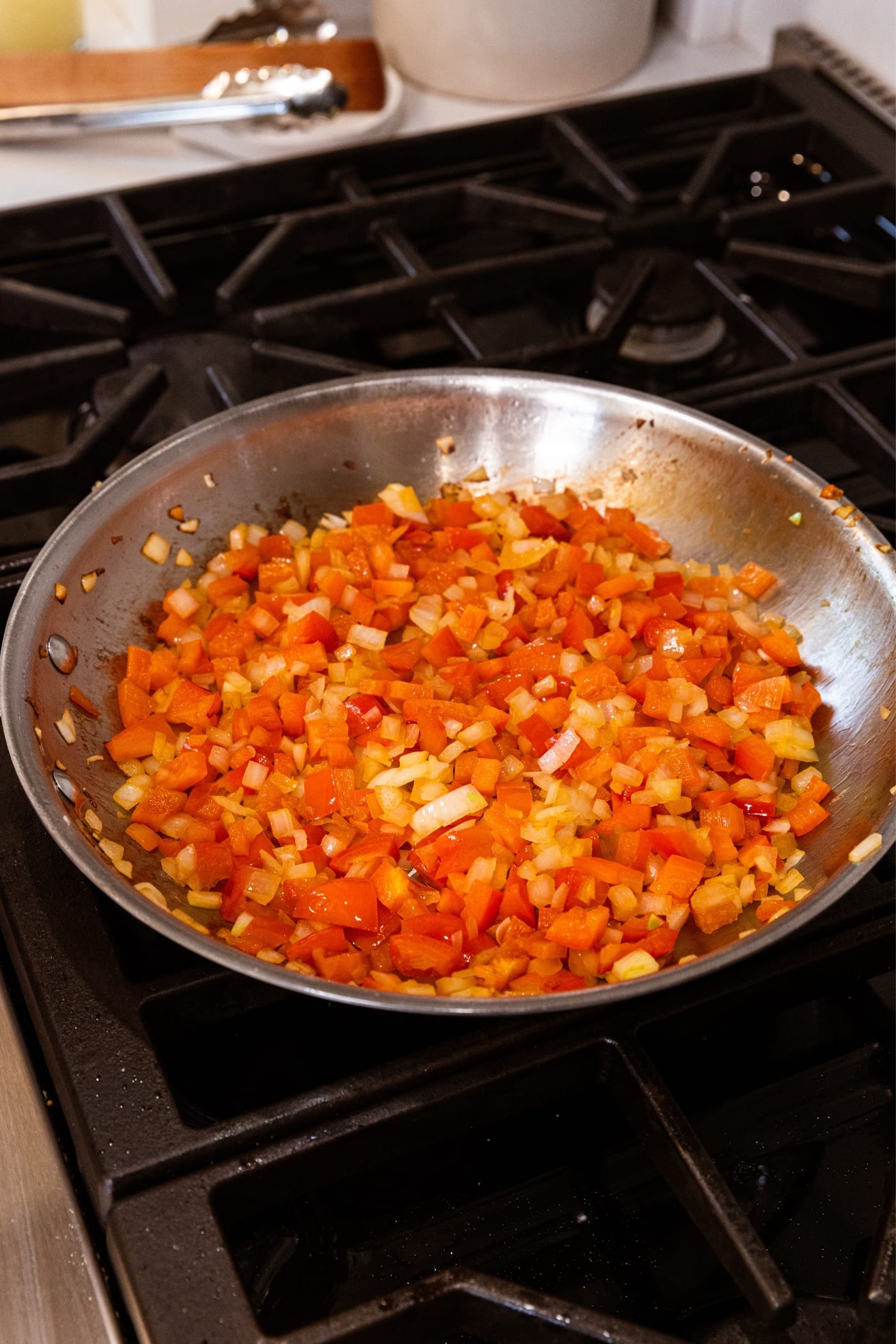 Metal skillet on stovetop filled with diced red bell pepper and onion over hot heat.