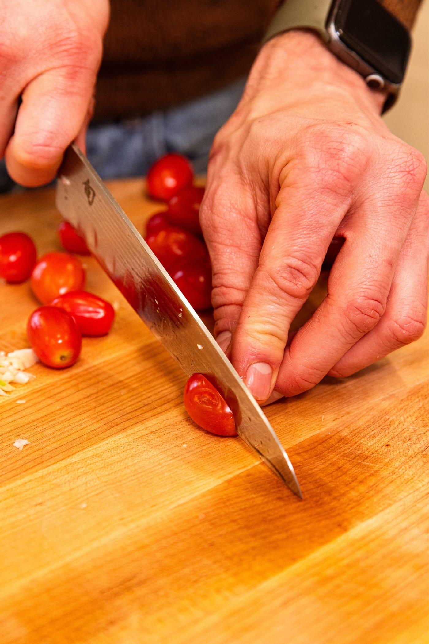 Hand using knife to slice cherry tomatoes in half on wooden cutting board.