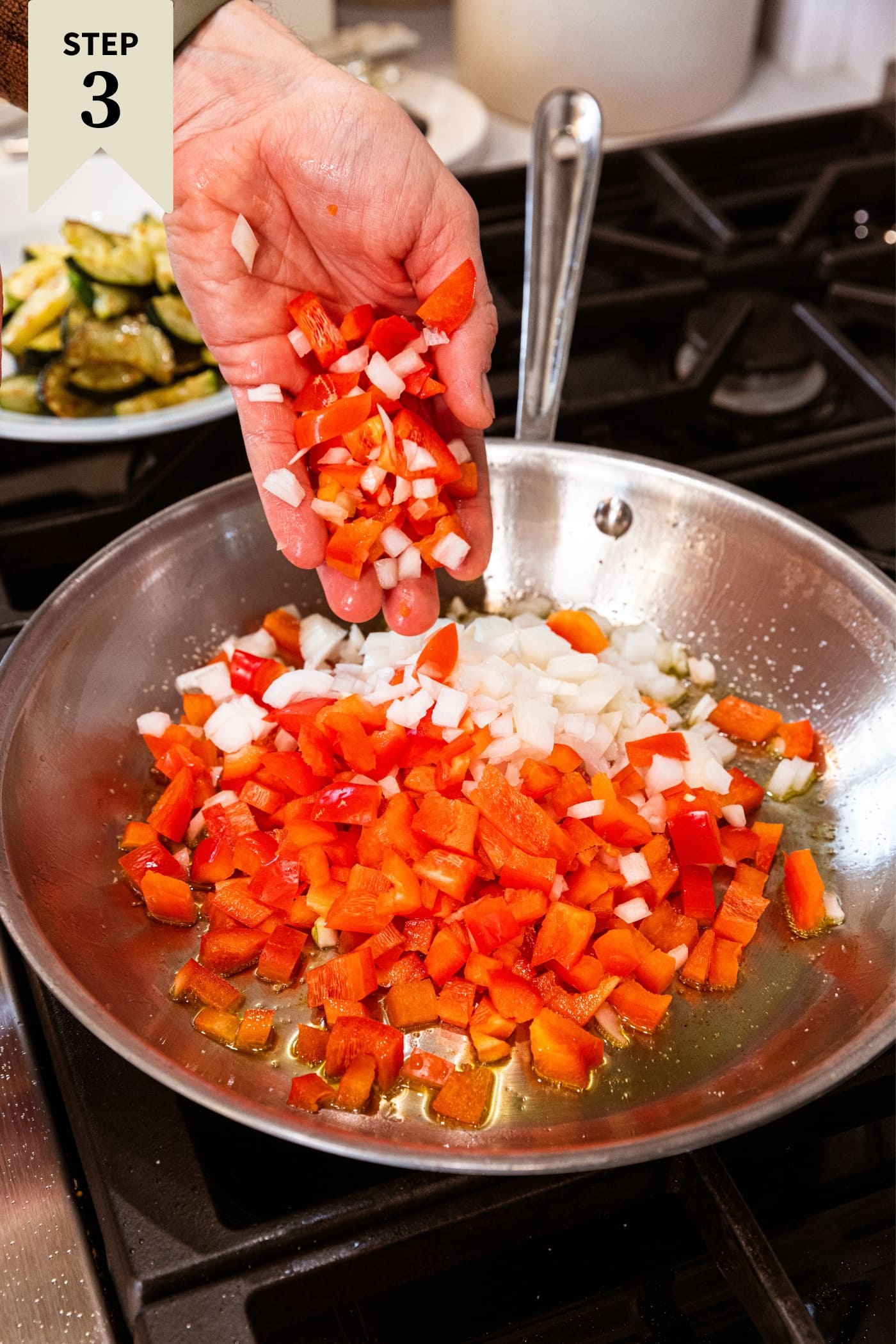Hand dropping diced red bell pepper and onion into a metal skillet on the stovetop with hot oil inside with cooked zucchini in background.