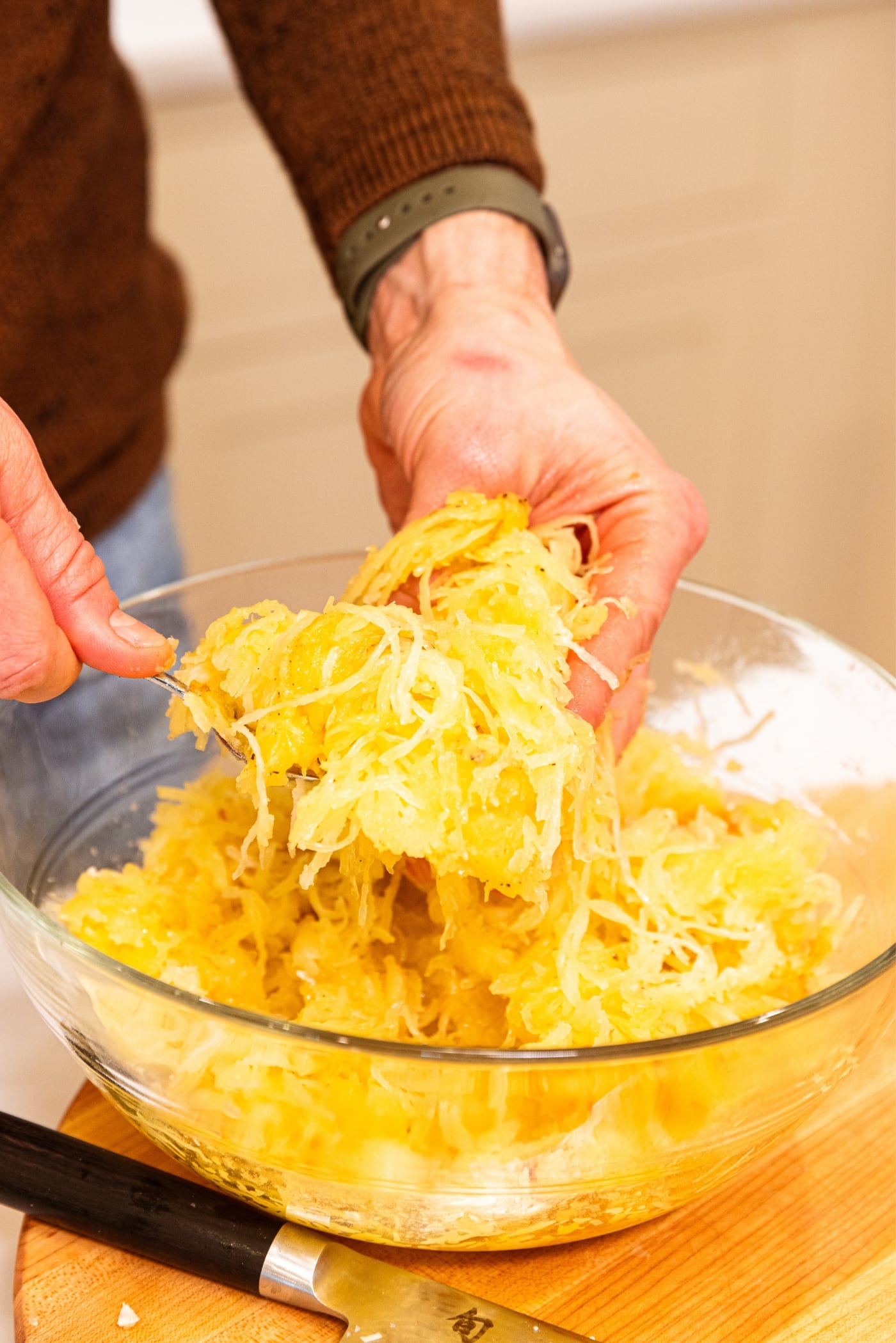 Hand using spoon and other hand to break up the texture of yellow-colored spaghetti squash in large glass bowl.
