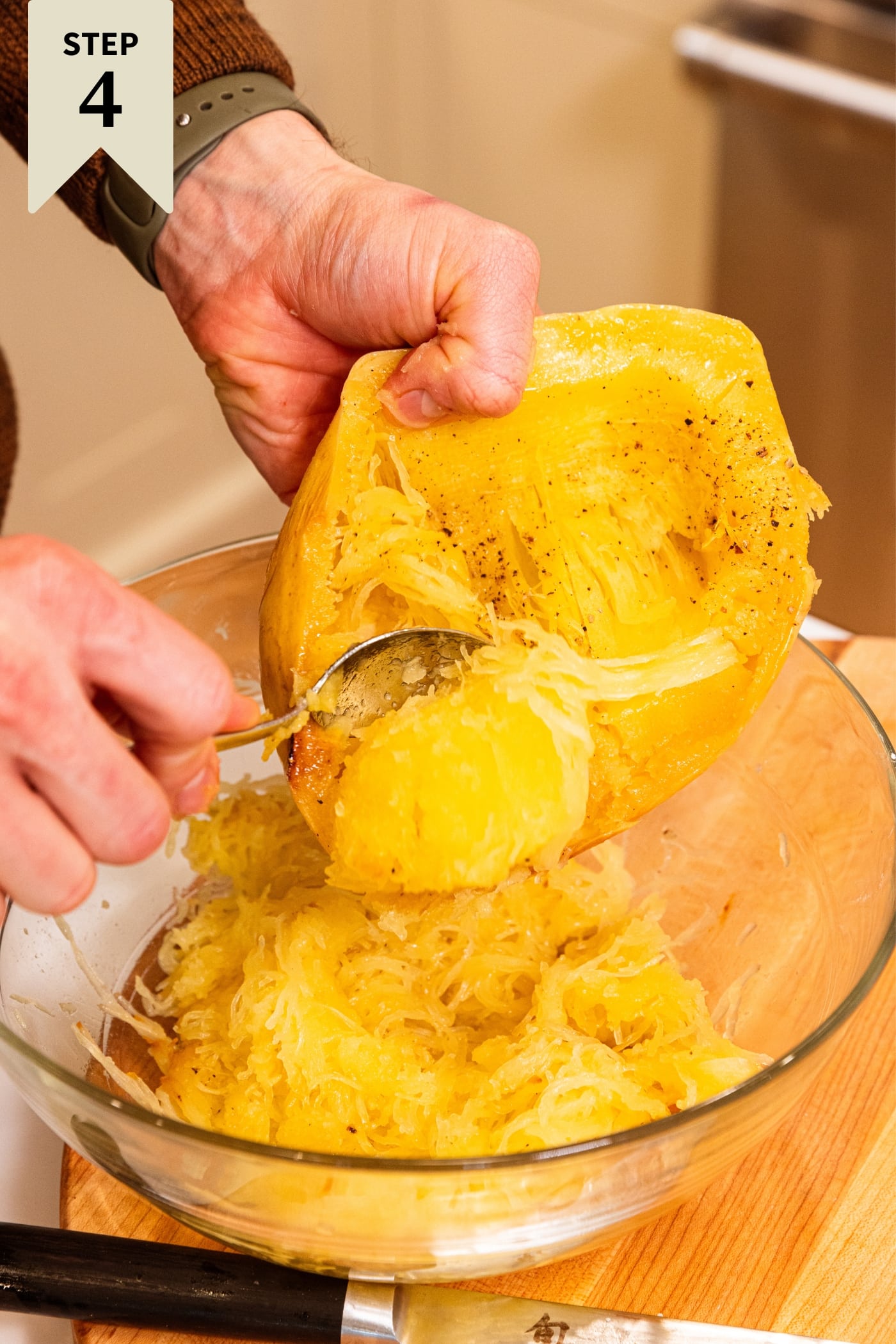 Hand holding roasted spaghetti squash scooping out the soft interior into a large glass bowl on wood cutting board.