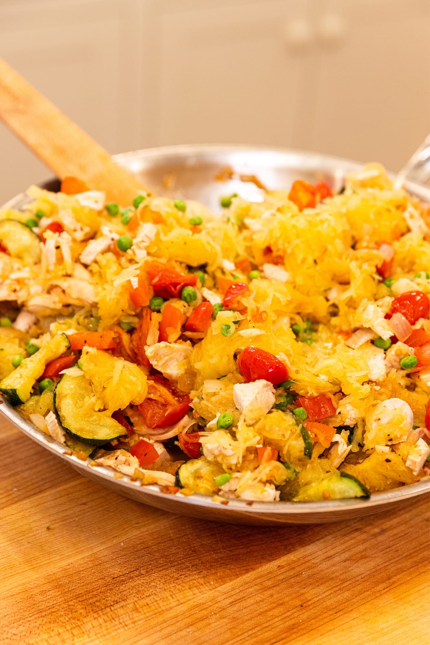 Metal skillet sitting on wooden cutting board filled with zucchini, tomatoes, chicken, and yellow-colored spaghetti squash after being mixed together.