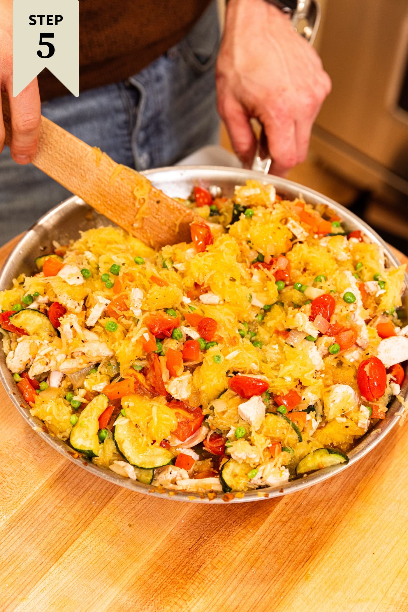 Metal skillet with tomatoes, spaghetti squash, chicken, and more vegetables inside being mixed together with wooden spatula on wooden surface.