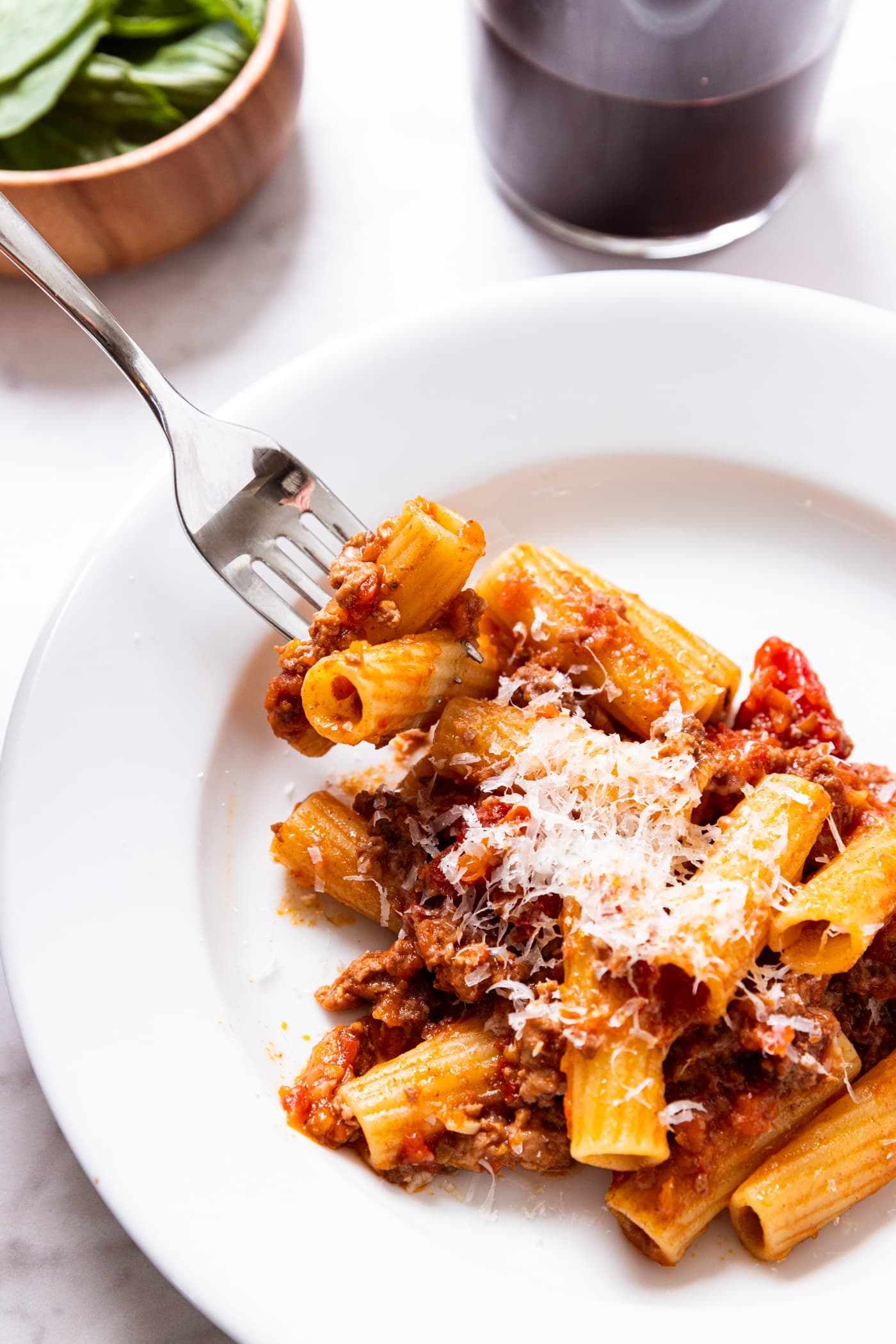 White plate with fork picking up tubes of pasta covered in bolognese sauce with small wooden bowl filled with basil and glass of red wine.