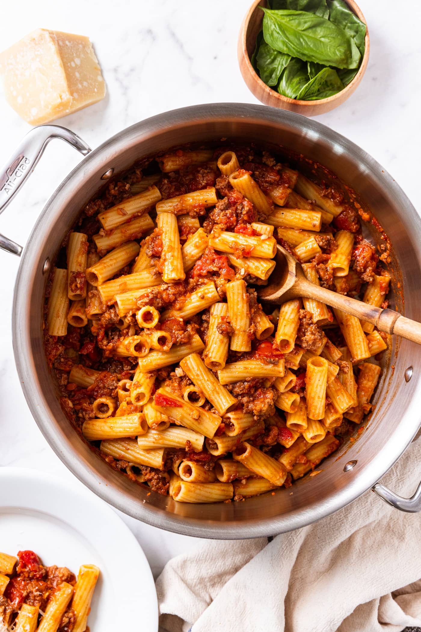White marble surface with large stockpot filled with cooked bolognese with wooden spoon resting in stockpot with basil in wooden bowl.