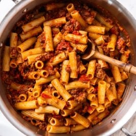 White marble surface with metal stockpot filled with cooked bolognese sauce tossed with pasta with wooden spoon resting on bottom of pan.