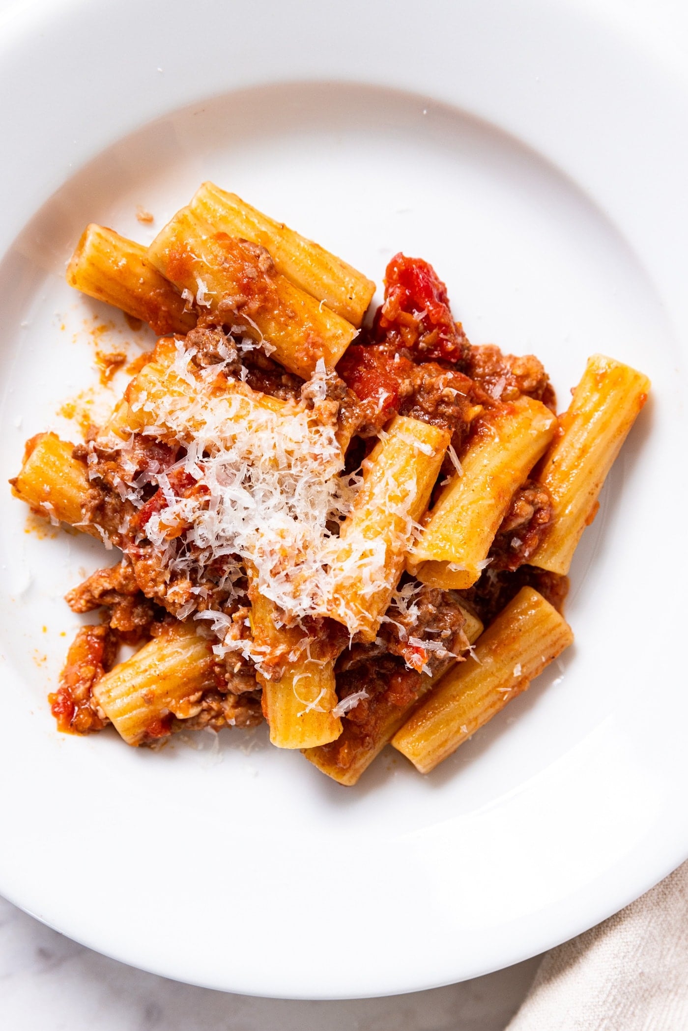 White plate filled with pasta topped with bolognese sauce and fresh shredded Parmesan cheese on top.