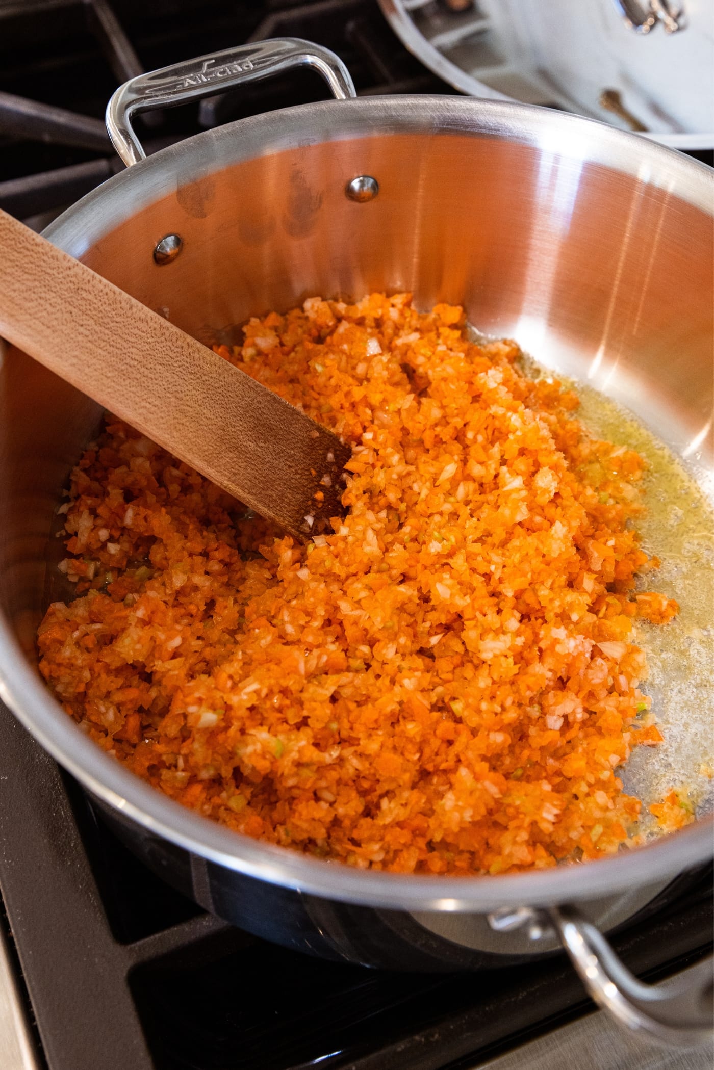 Wooden scraper mixing around roughly chopped carrots, onion, and celery in melted butter in metal stockpot on stovetop.