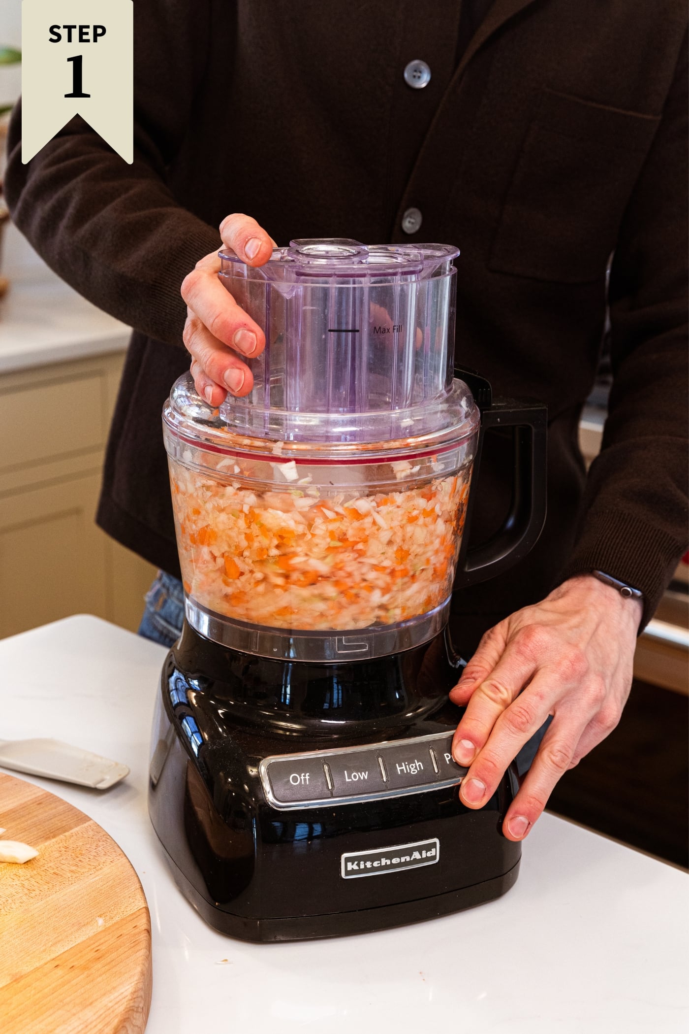 Hand pushing button on food processor to begin blending roughly chopped pieces of onion, carrot, and celery on white surface.