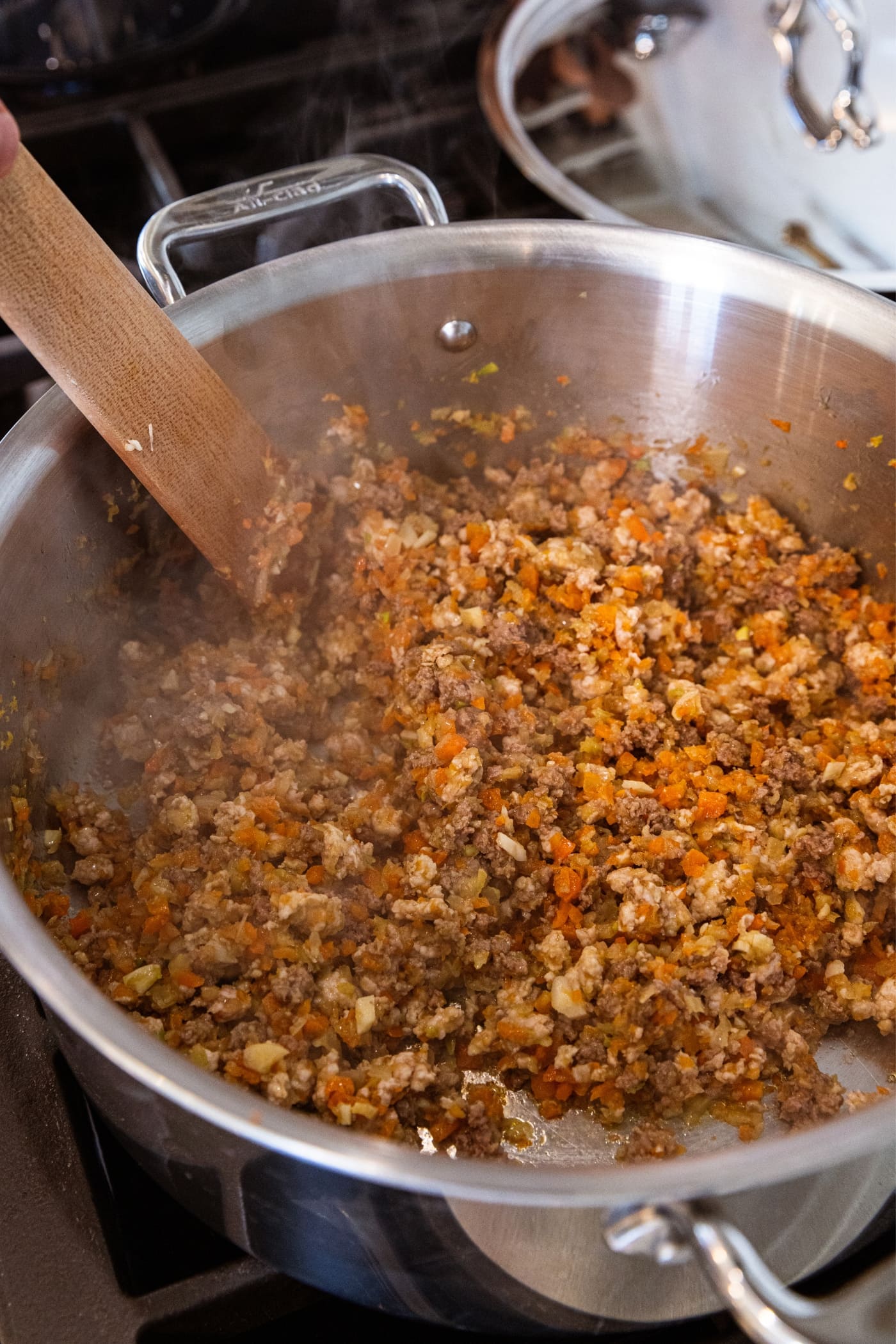 Browned meat and carrot, onion, and celery mixture sitting in bottom of metal stockpot on the stove with wooden scraper resting on side of pan.