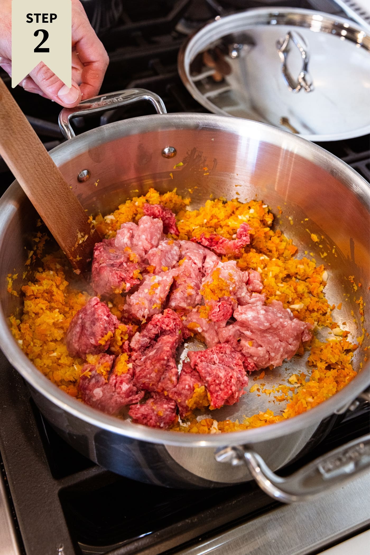 Metal stockpot sitting on stovetop with raw meat in bottom along with chopped carrots, onion, and celery, all about to be browned with melted butter.