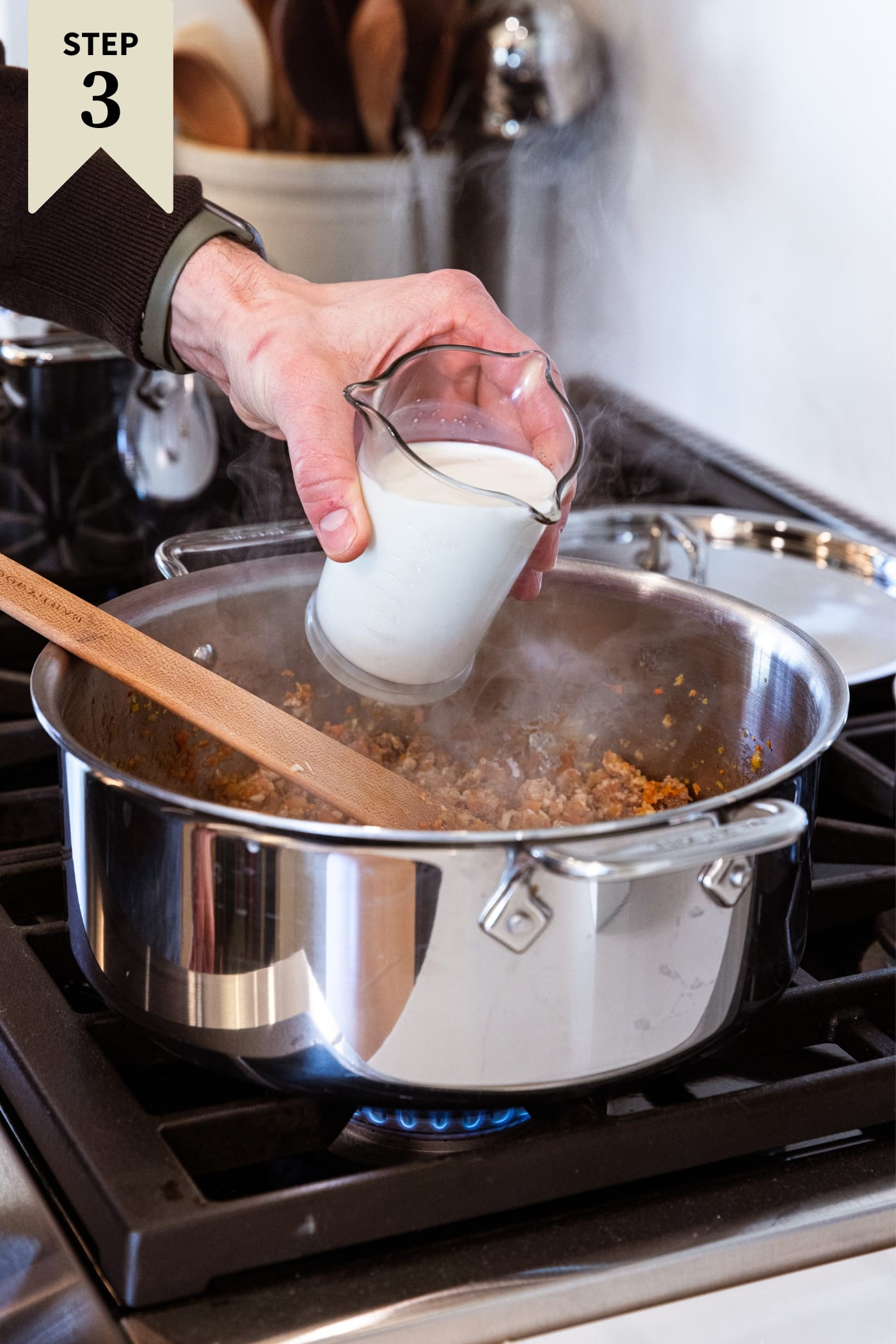 Hand holding measuring cup full of whole milk about to pour it into a large metal stockpot on the stovetop.