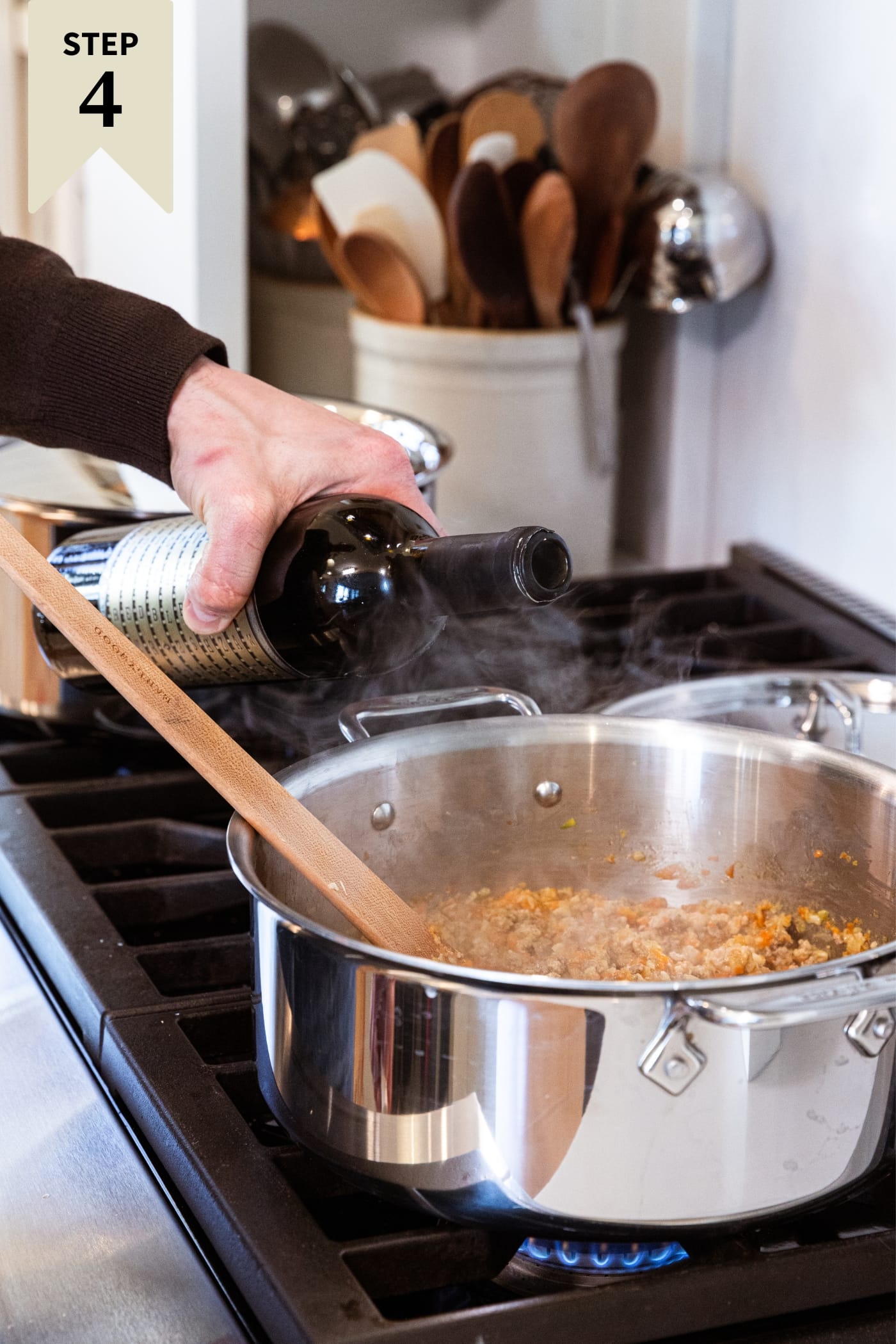 Hand holding bottle of red wine about to pour it into a large metal stockpot on the stove filled with meat and vegetables cooking.