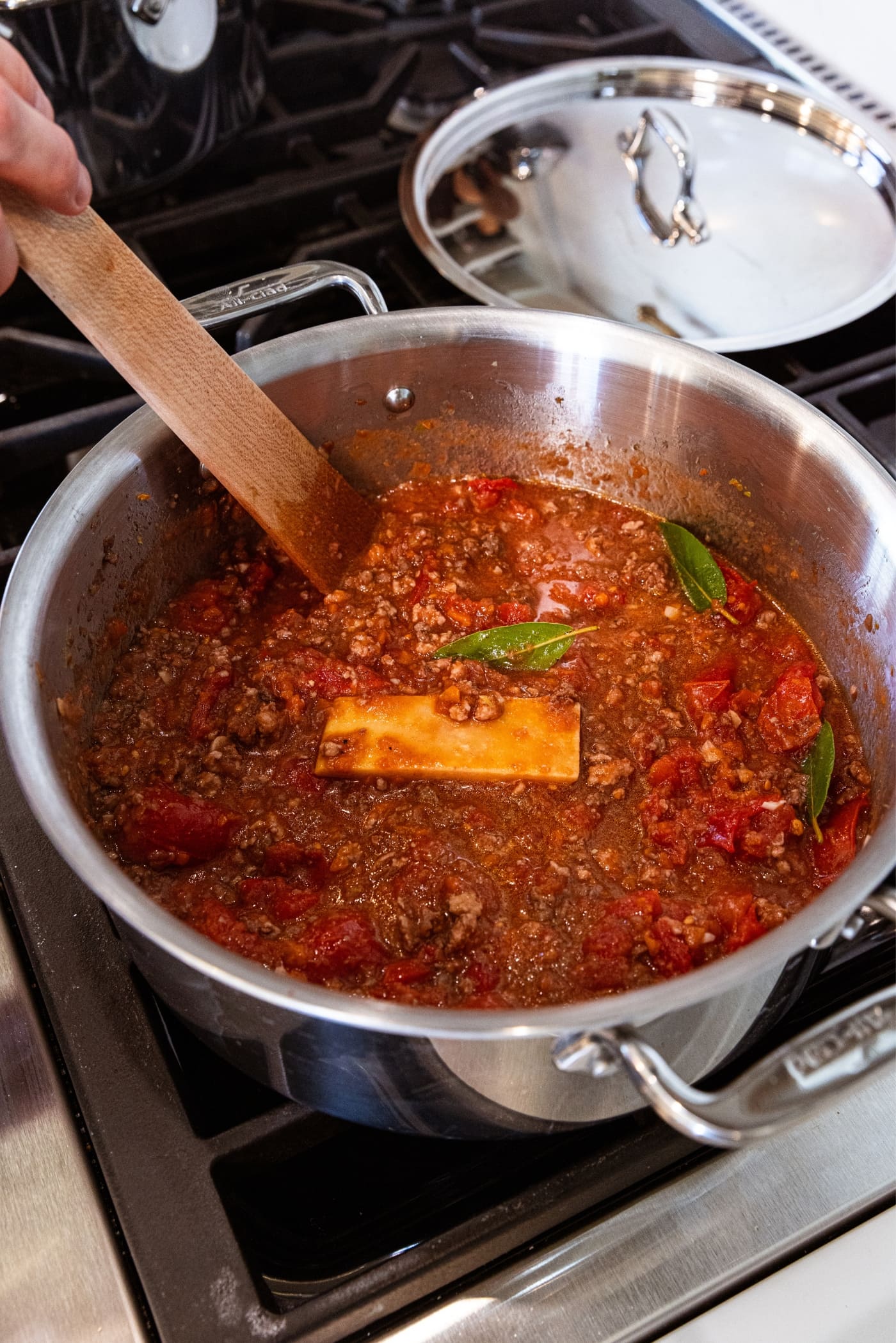 Large metal stockpot filled with tomatoes along with meat and vegetables with Parmesan rind and bay leaves sitting on the surface.