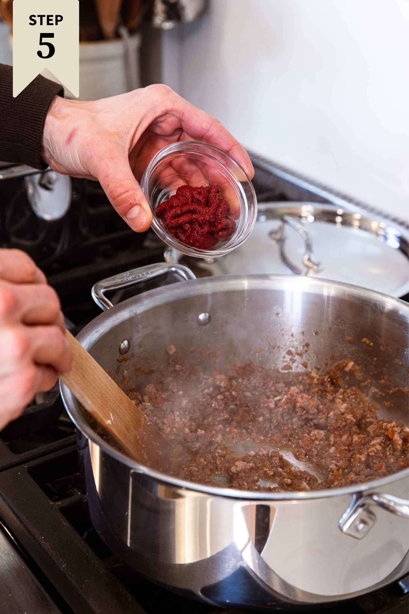 Hand holding small glass bowl with tomato paste inside before pouring it into a simmering mixture of meat and vegetables.