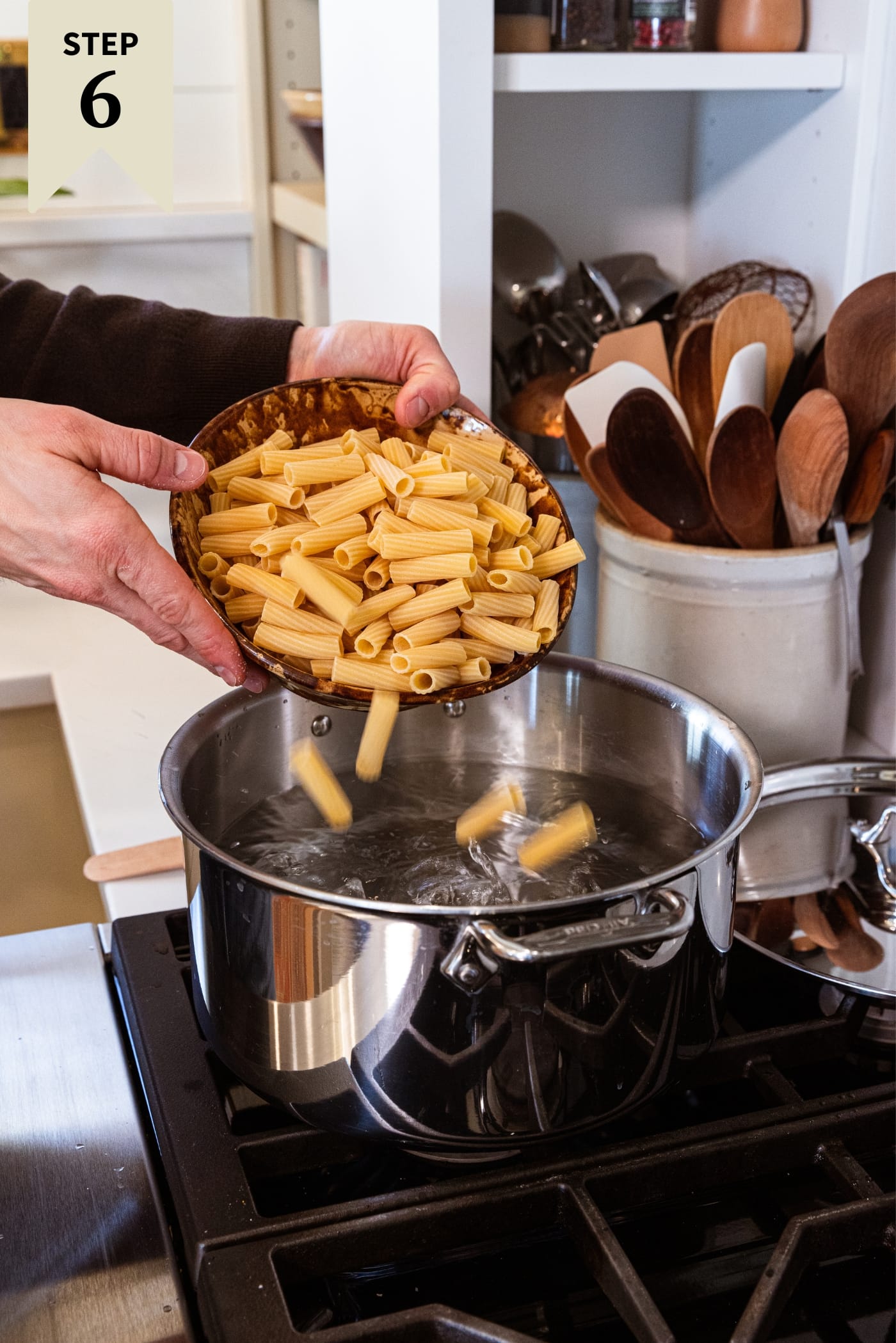 Hands holding brownware bowl filled with pasta pouring into a large stockpot filled with water.