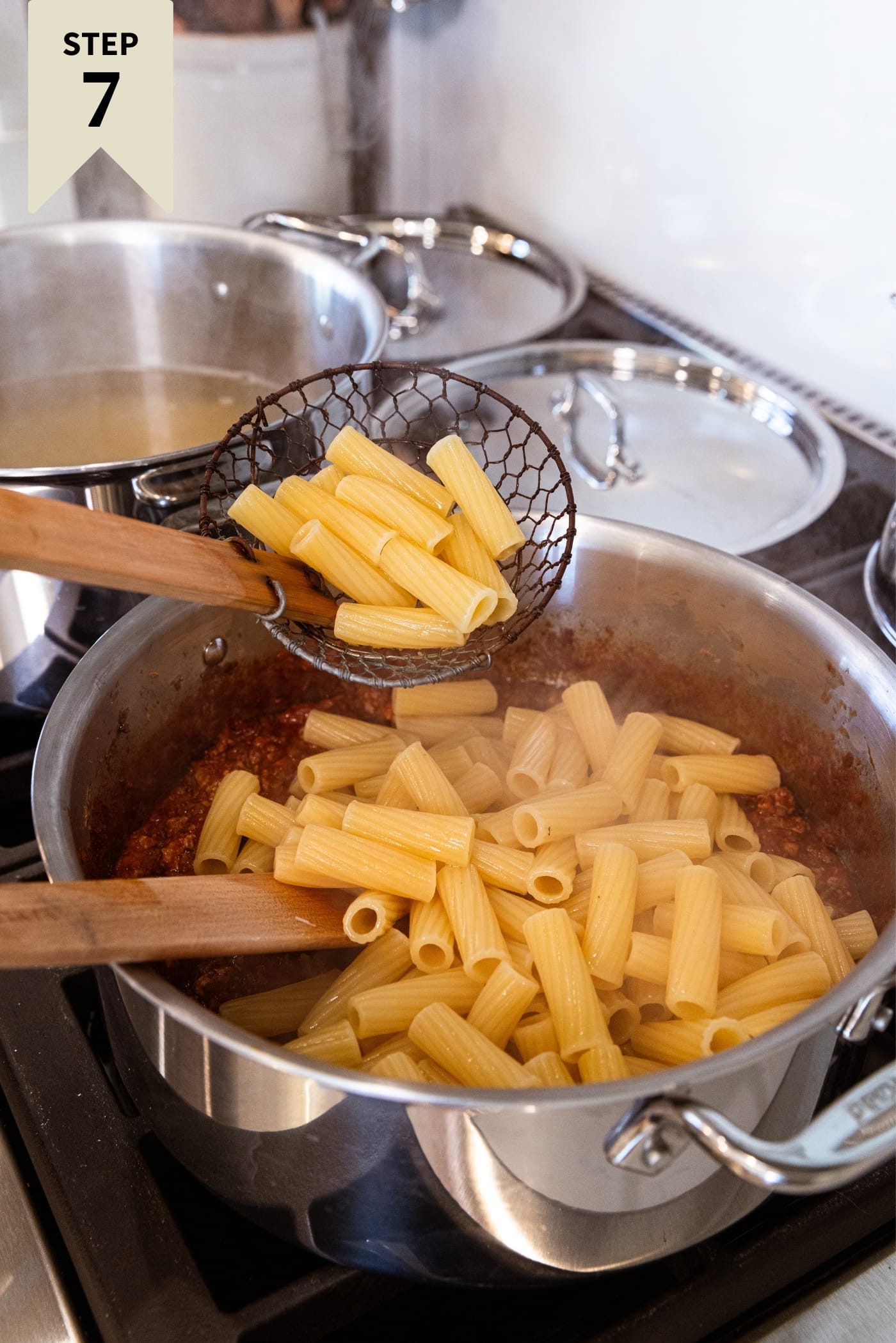Spider pouring cooked pasta into a large metal stockpot filled with more pasta and tomato meat sauce before being mixed together.