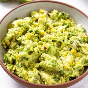 White marble surface with brown bowl filled with sweet corn guacamole with chunky texture.