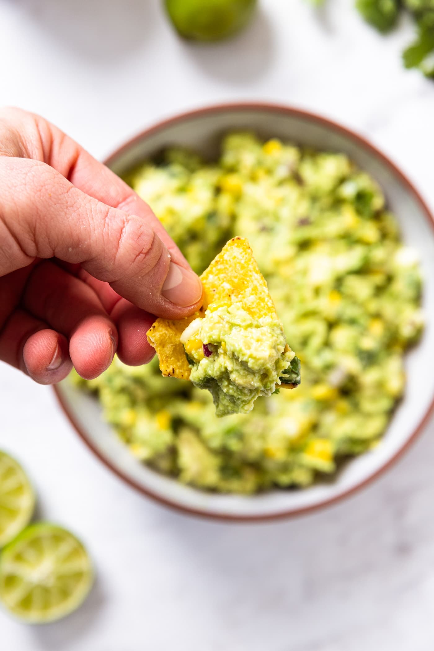 Hand holding corn tortilla chip with scoop of guacamole with rest of guacamole in bowl below on white surface.