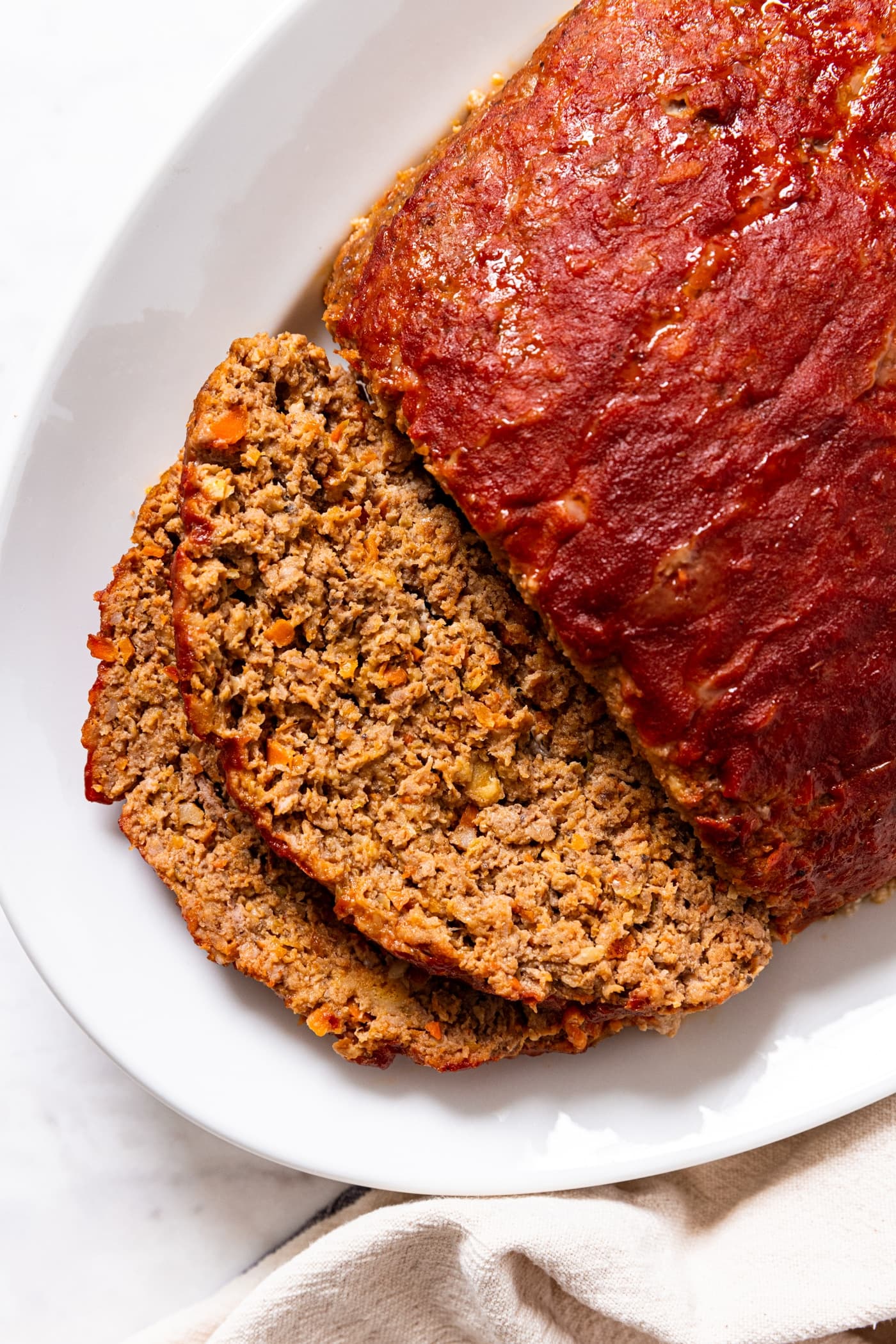 White serving platter with meatloaf sitting with two slices sliced off the front with ketchup slathered all over the top showing interior texture of meat.