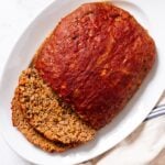 White platter sitting on white marble surface filled with meatloaf with two slices sliced off the side with ketchup all over the top.